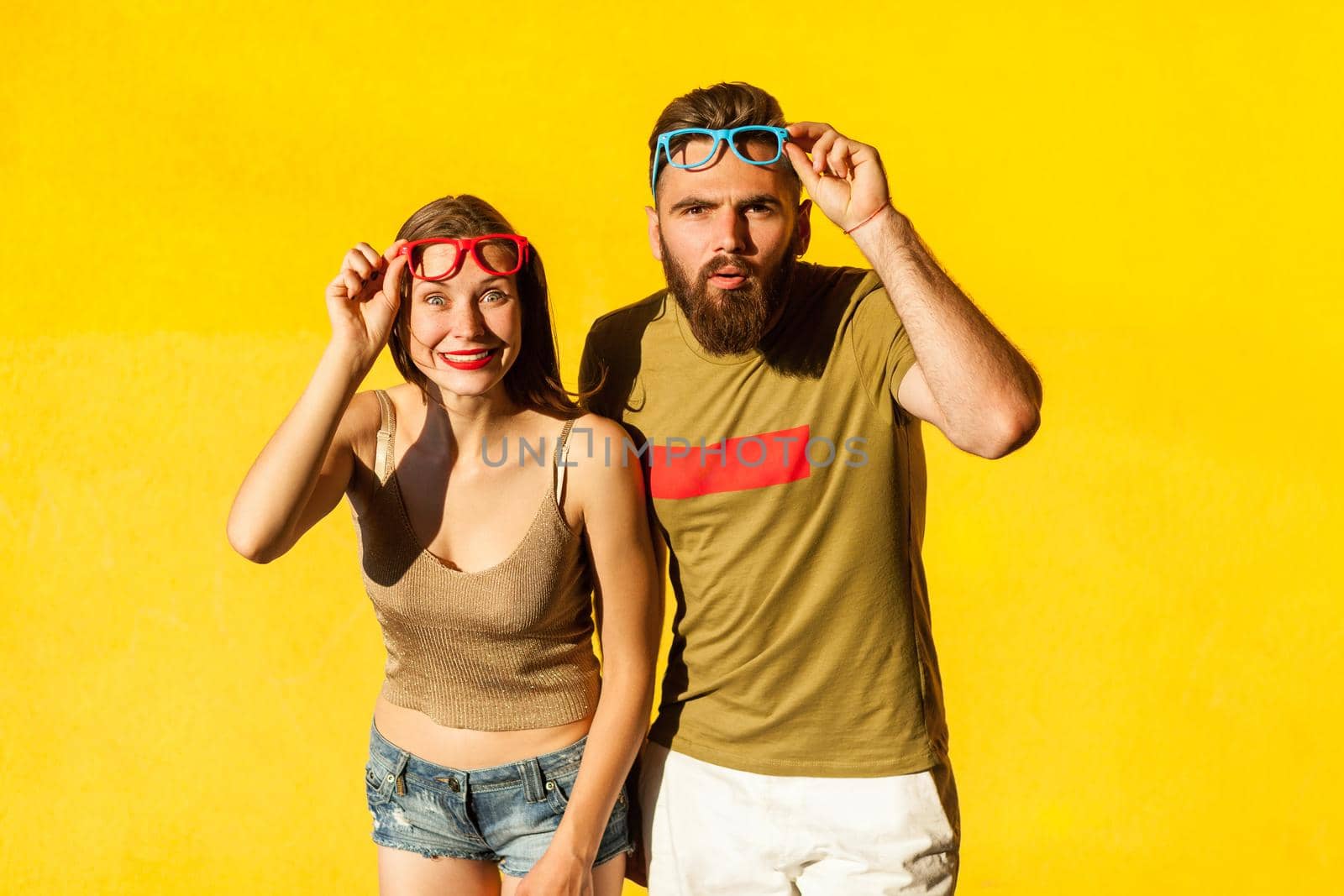 Funny couple in casual style clothes and color glasses looking at camera. Isolated on yellow background , indoor studio shot
