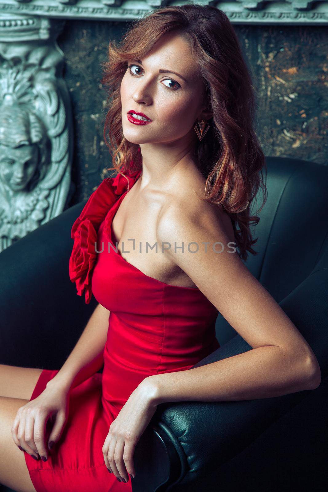Beautiful young fashion model in red dress and fashion makeup and hairstyle posing sitting in the armchair near the artificial fireplace and looking at camera. Studio shot.
