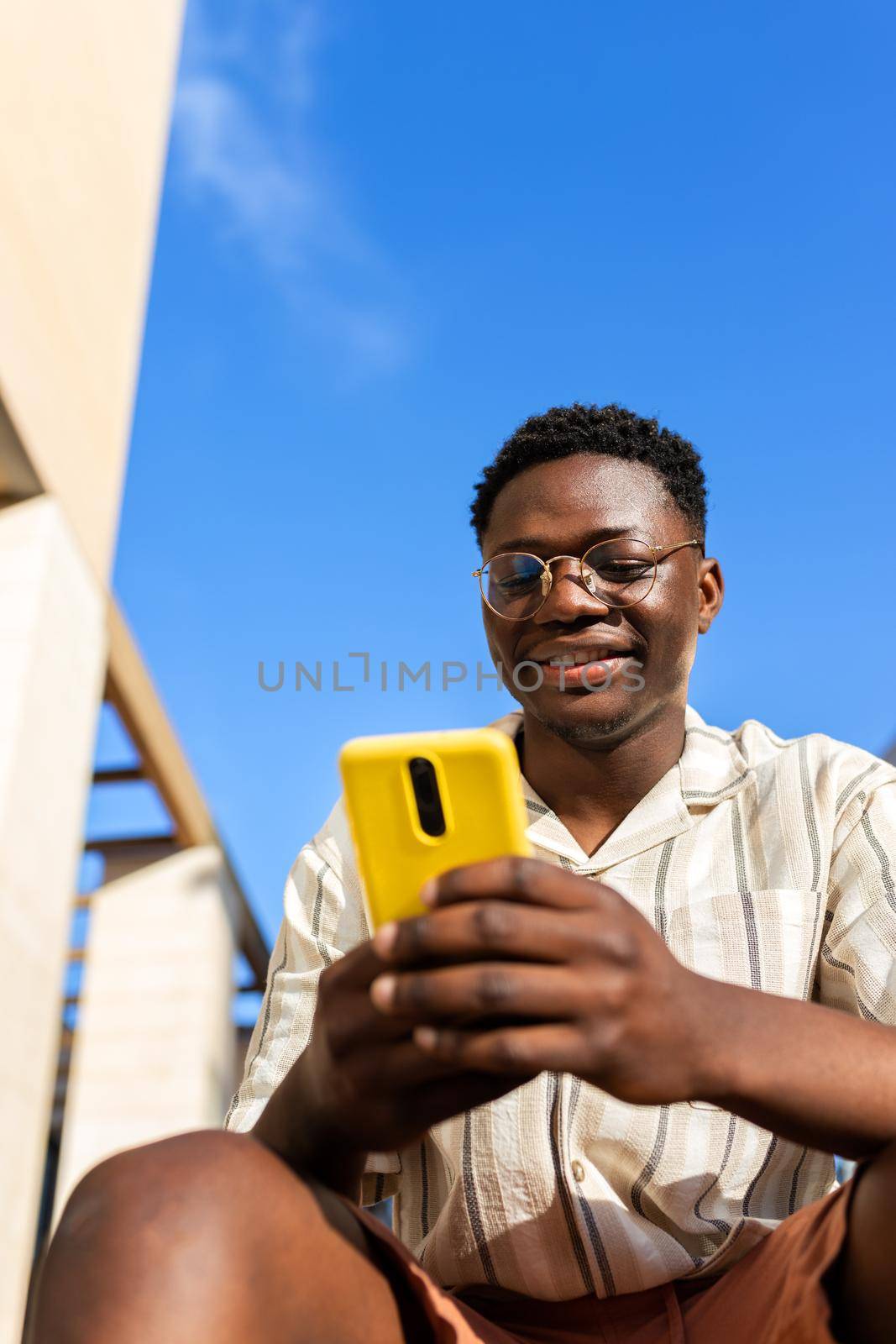 Black young man sitting on stairs outdoors using mobile phone. Vertical image. Copy space. by Hoverstock