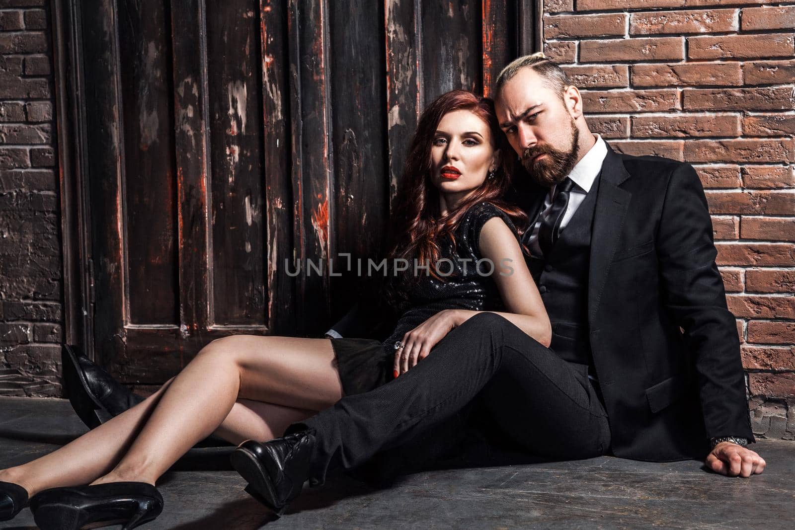 Fashion studio photo of sexy couple of ginger woman and blonde man dressed wearing classic style, sitting on the flor. Isolated on brick wall, studio shot.