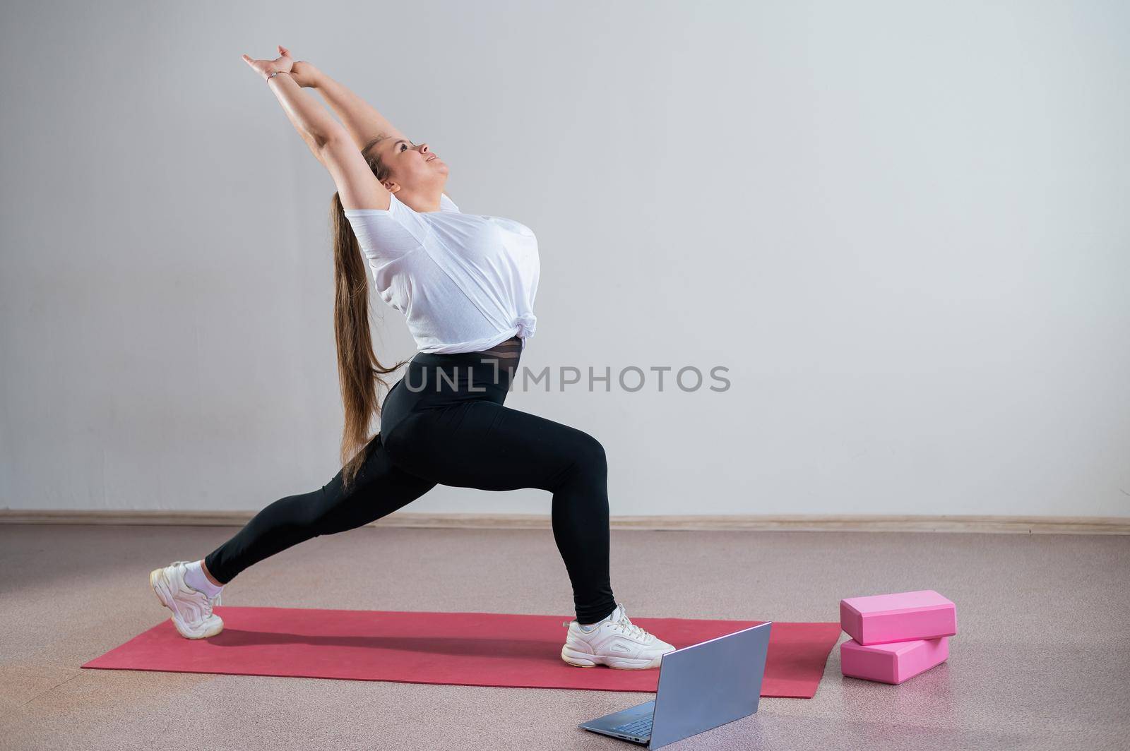 Young Plus Size Woman Stretching At Home Online. Flexible girl practices yoga and watches an online course on a laptop.