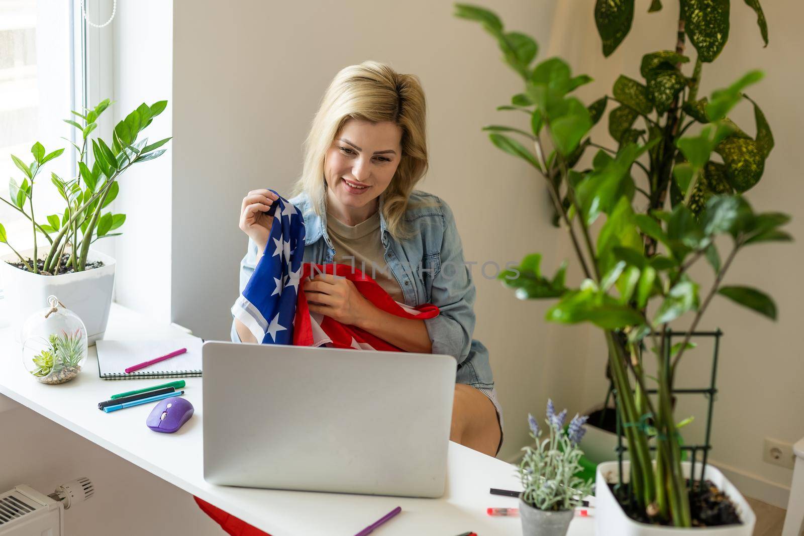 Happy young female student hold flag of USA, studying with laptop with blank screen in living room interior. International education at home, lesson remote, website due covid-19 quarantine by Andelov13