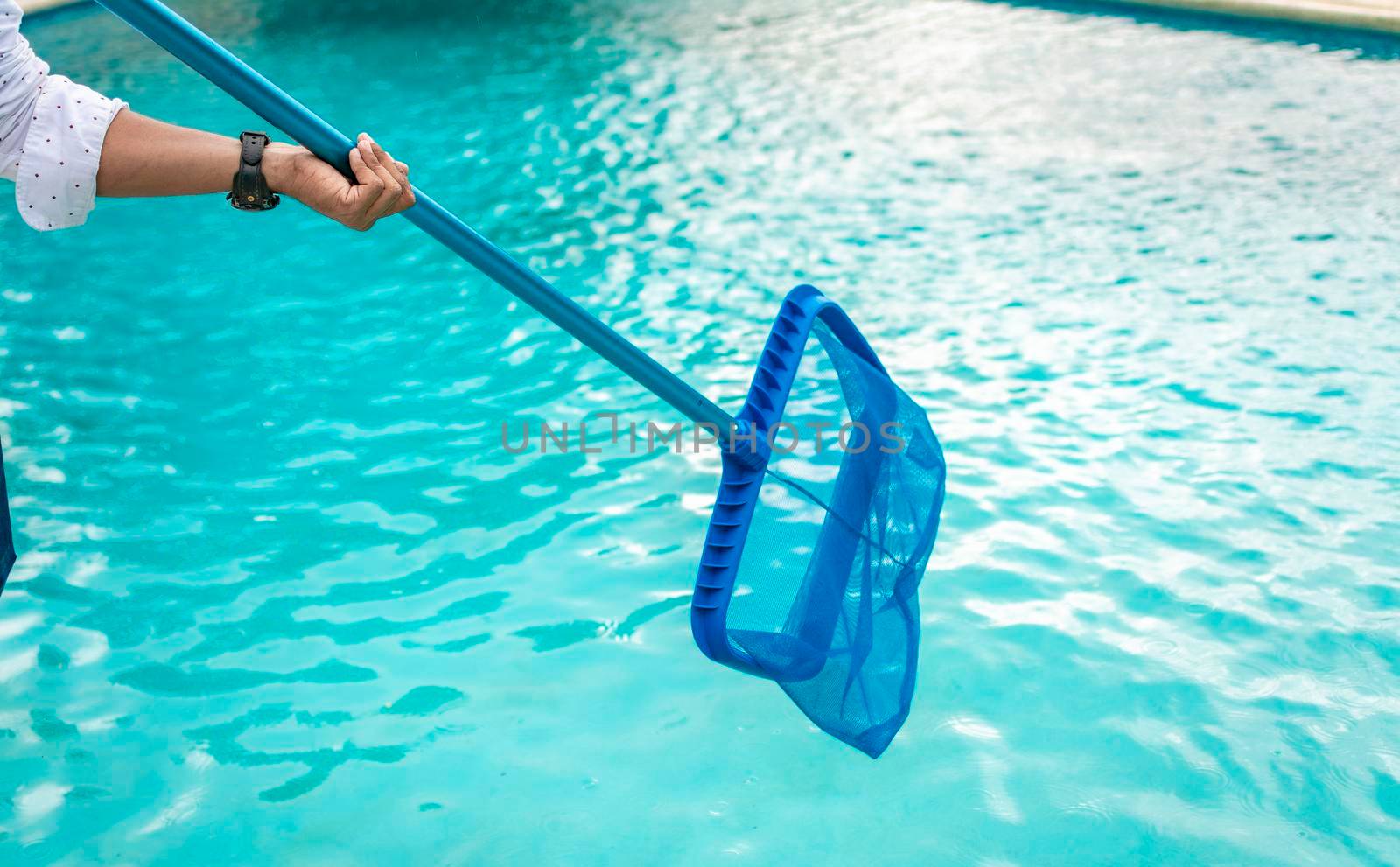 A man cleaning pool with leaf skimmer. Man cleaning the pool with the Skimmer, Person with skimmer cleaning pool, Hands holding a skimmer with blue pool in the background