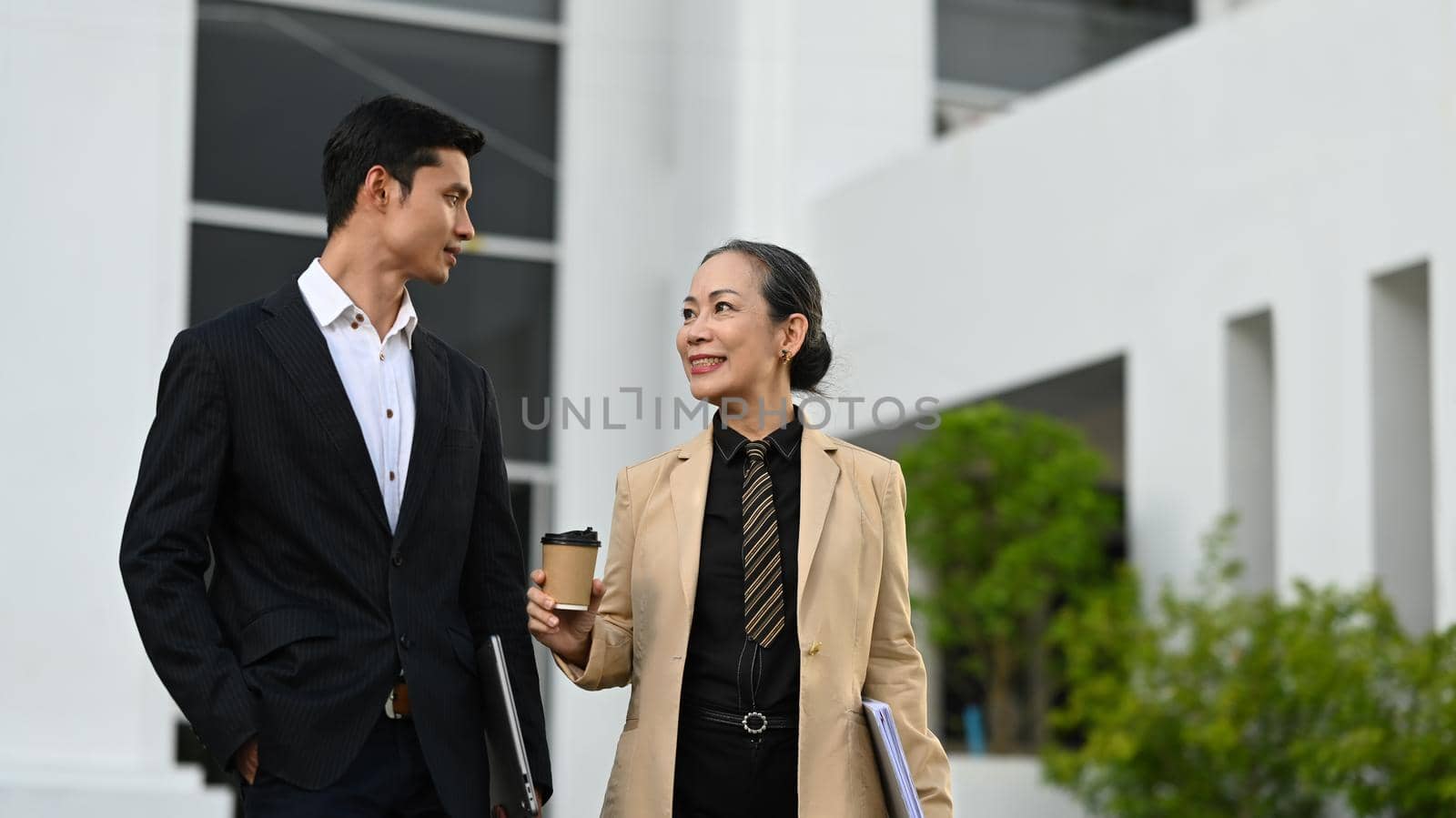 Two happy business people wearing suits walking outside office building and talking each other.