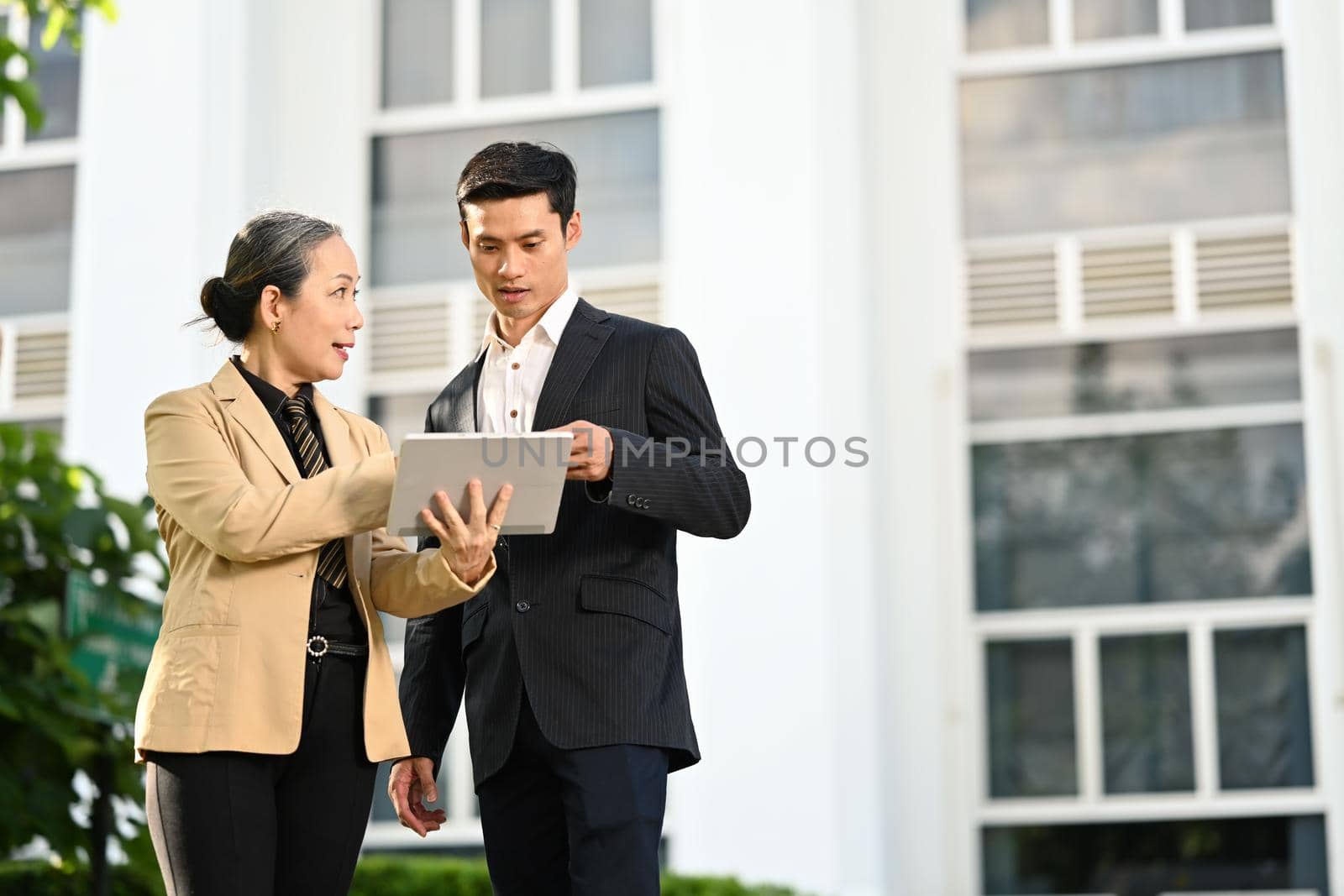 Senior female entrepreneur and young businessman discussing work on tablet while standing outdoor with modern office building in background by prathanchorruangsak