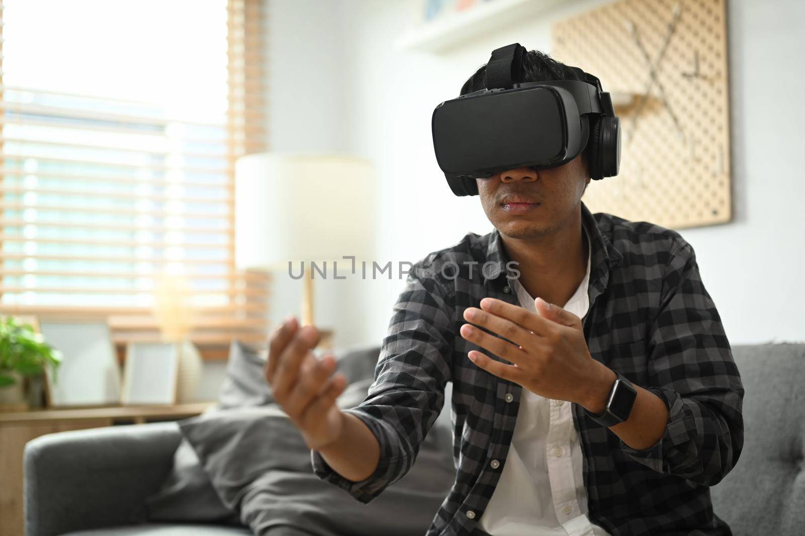 Young asian man sitting on couch and playing video games in VR glass headset.