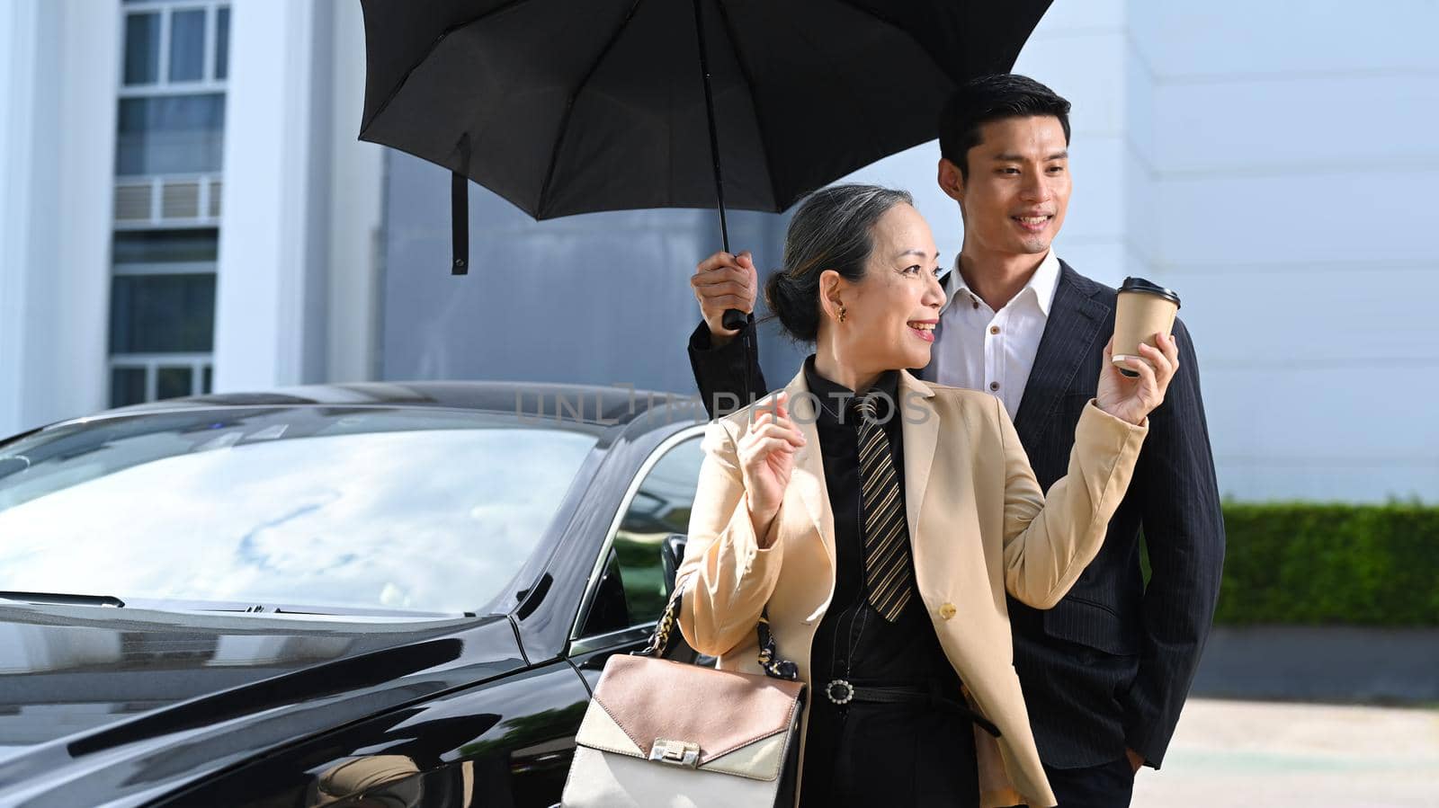 Smiling mature businesswoman and her assistant standing by black car at parking with modern office building in background by prathanchorruangsak