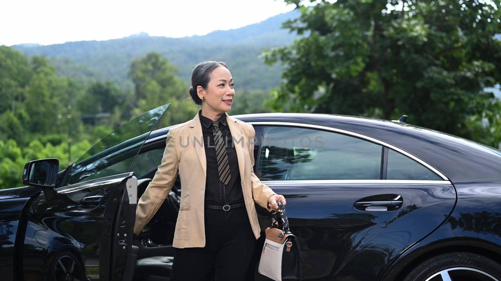 Elegant mature businesswoman standing by open door of black car and looking alway.