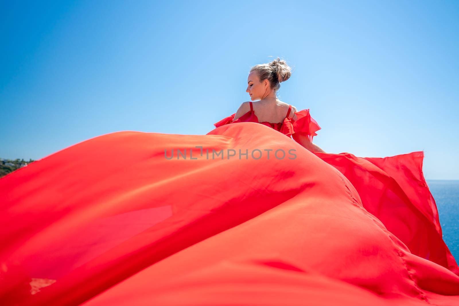 Blonde with long hair on a sunny seashore in a red flowing dress, back view, silk fabric waving in the wind. Against the backdrop of the blue sky and mountains on the seashore. by Matiunina