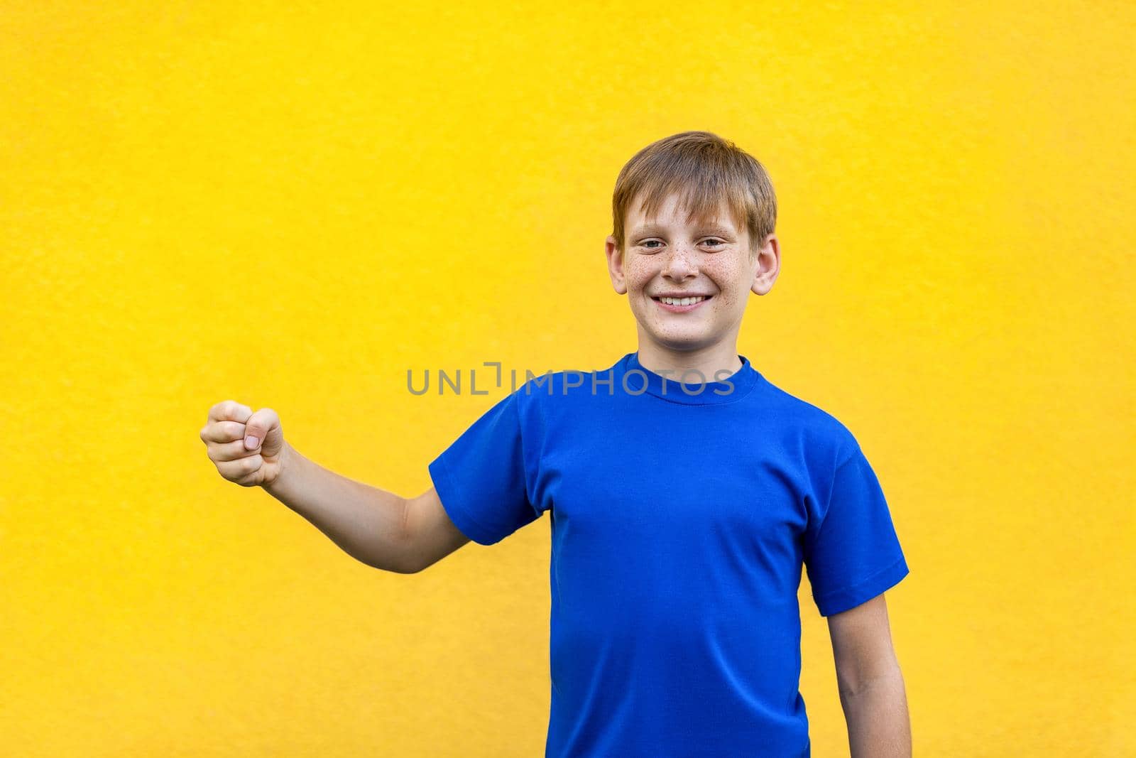 Happiness freckled boy holding imaginary object, looking at camera with toothy smile. Studio shot, yellow background