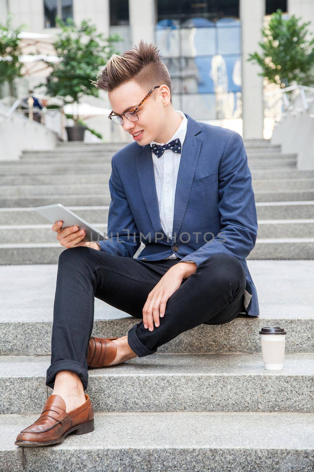 Attractive young businessman sitting with tablet and coffee in hands on office building background. holding tablet and looking at display with toothy smile.