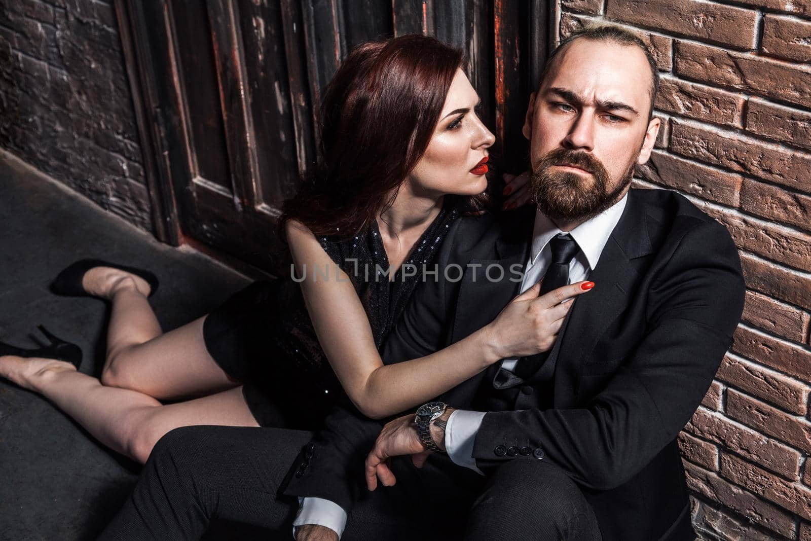 Man in suit and woman in black evening dress sitting on his lap. Foxy woman looks at him and wants to kiss. Studio shot, isolated on brick background