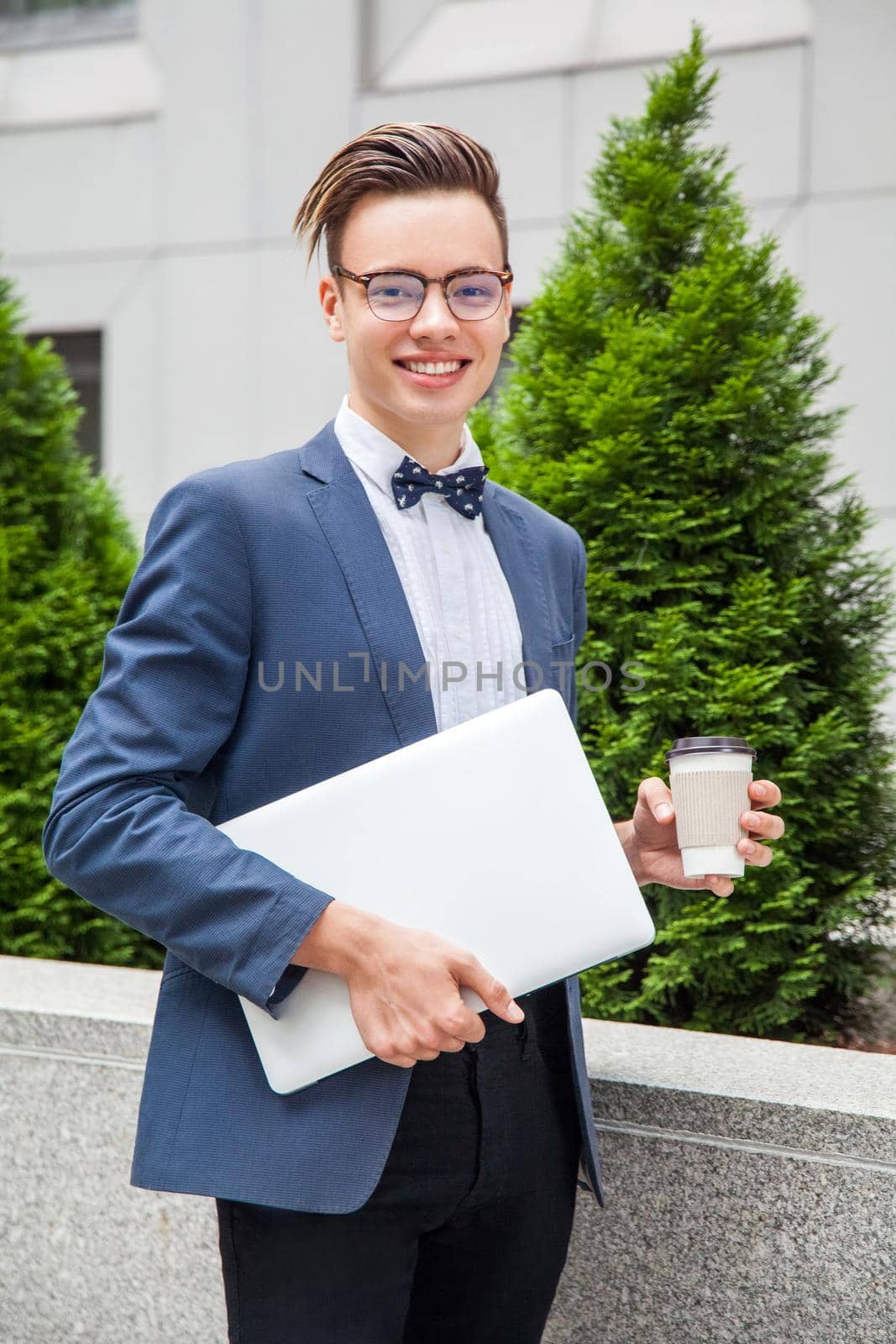 Attractive young businessman with laptop and coffee in hands on office building background. holding laptop and coffee and looking at camera with toothy smile.
