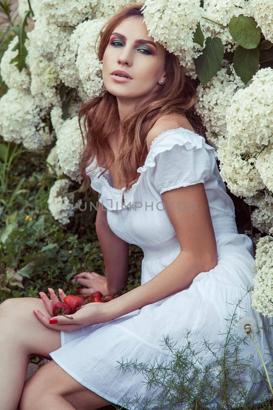 Woman sitting in park near many flowers ,holding strawberry. Outdoor spring or summer photo