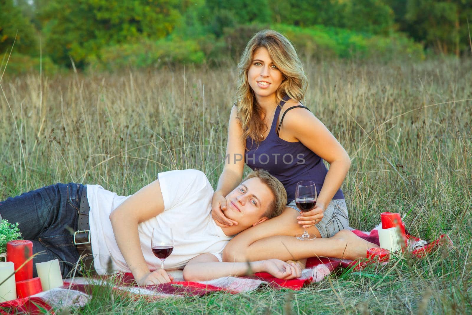 Beautiful young happy loving couple on picnic lying down on plaid in field on sunny summer day enjoying, holding and drinking wine and resting. looking at camera and smiling.