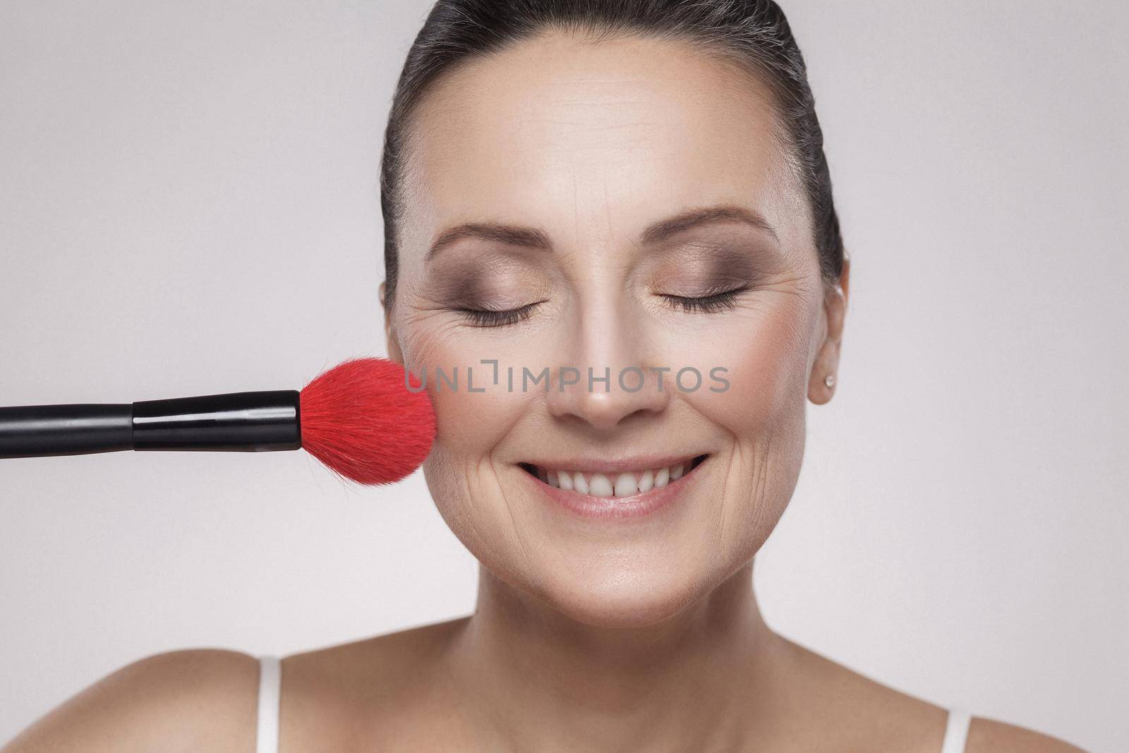 Closeup portrait of a attractive middle aged woman with perfect skin applying makeup with a red brush and toothy smile against a gray background. Indoor, studio shot, copy space, looking at camera