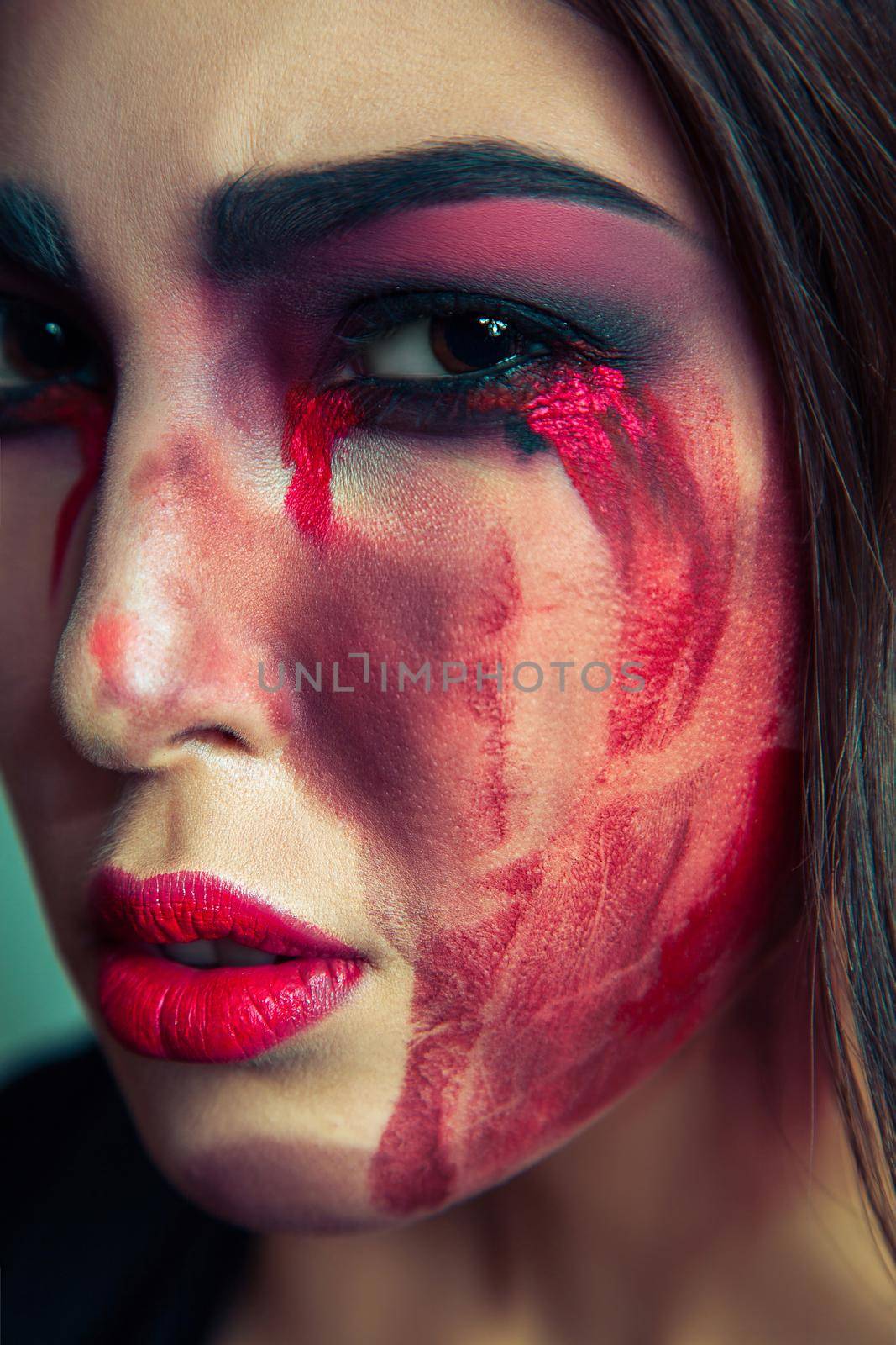 portrait of freak monster with mess dirty colored makeup on her face. crying woman with red bloody tears and hand. halloween concept on green background. studio shot, dark brown eyes.