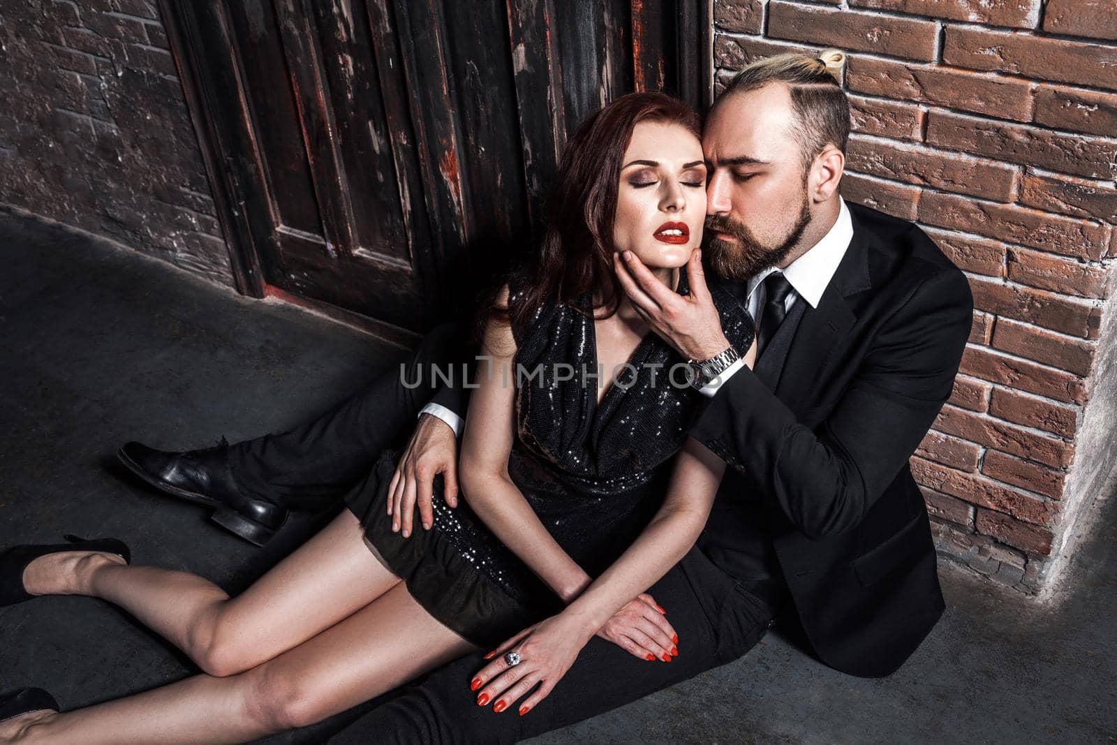 Handsome bearded man sitting on the floor, touching chin woman, while his ginger girlfriend is leaning on him. Studio shot, vintage inerior