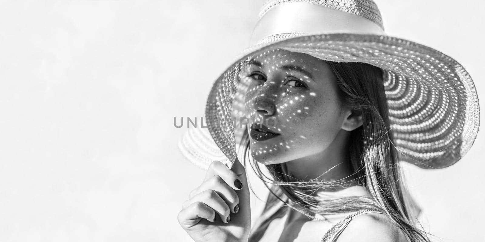 Portrait of beautiful sensual brunette young woman with makeup looking at camera with passion, holding hat, light through hat falls on face. Indoor studio shot isolated on light background copyspace.