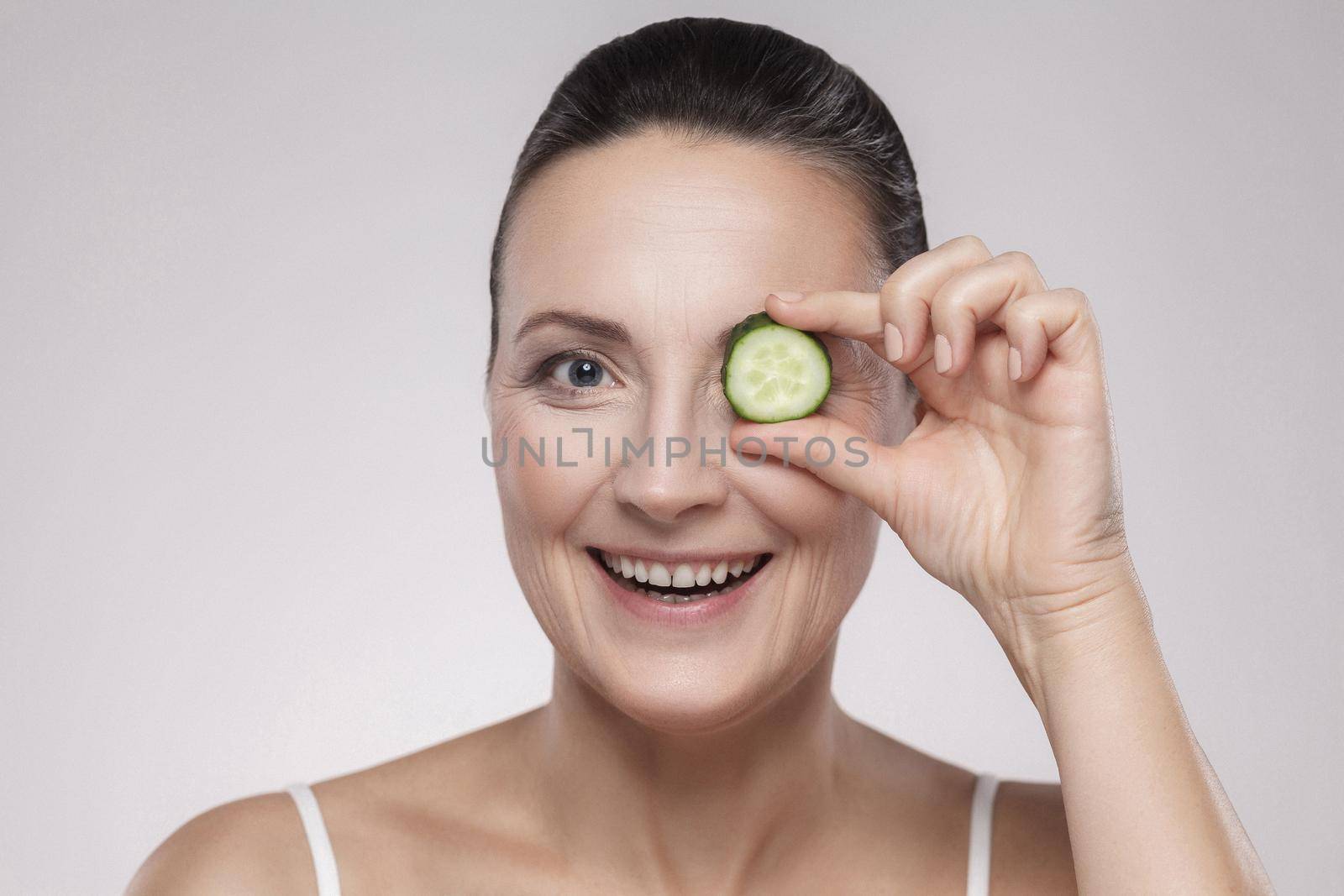 Sensitive care for luminous skin. Attractive middle aged woman looking at camera and covering eye with slice of cucumber while standing against grey background. Copy space, indoor,studio shot,isolated
