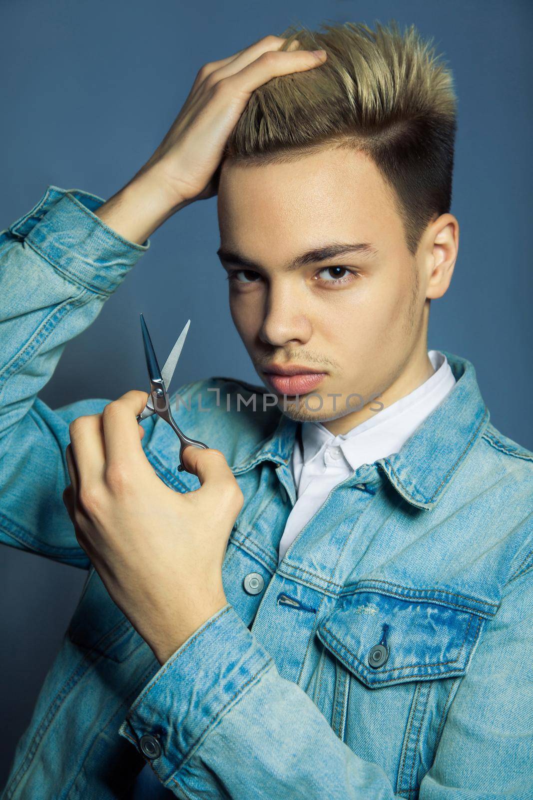 Portrait of fashion blonde Barber hairstylist with scissors in hand near eye on blue background.