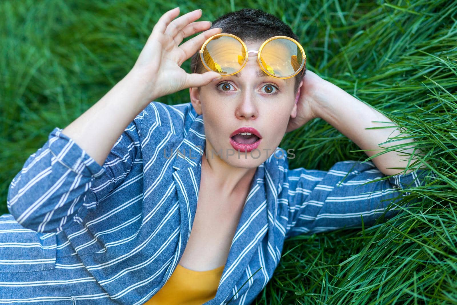 Portrait of surprised cute young woman with short hair in casual blue striped suit lying down on green grass, hoding yellow glasses up and looking at camera with amazed face. outdoor summertime shot.