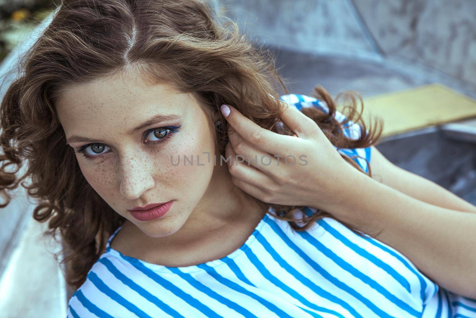 Beautiful young fashion model with freckles on her face and light blue striped dress and fashion makeup and hairstyle is lying down in metalic silver boat on lake, posing and looking at camera with blue eyes.