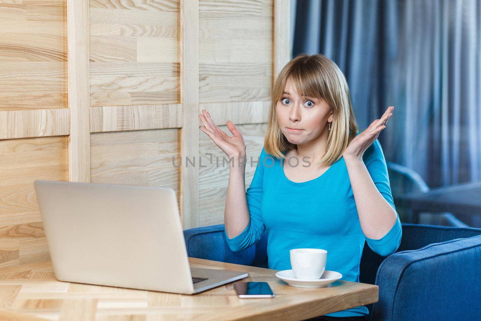 Not sure! Portrait of confused young girl freelancer with blonde bob haircut hair in blue t-shirt are sitting in cafe and working on laptop with raised arms and puzzled face, looking at camera. indoor