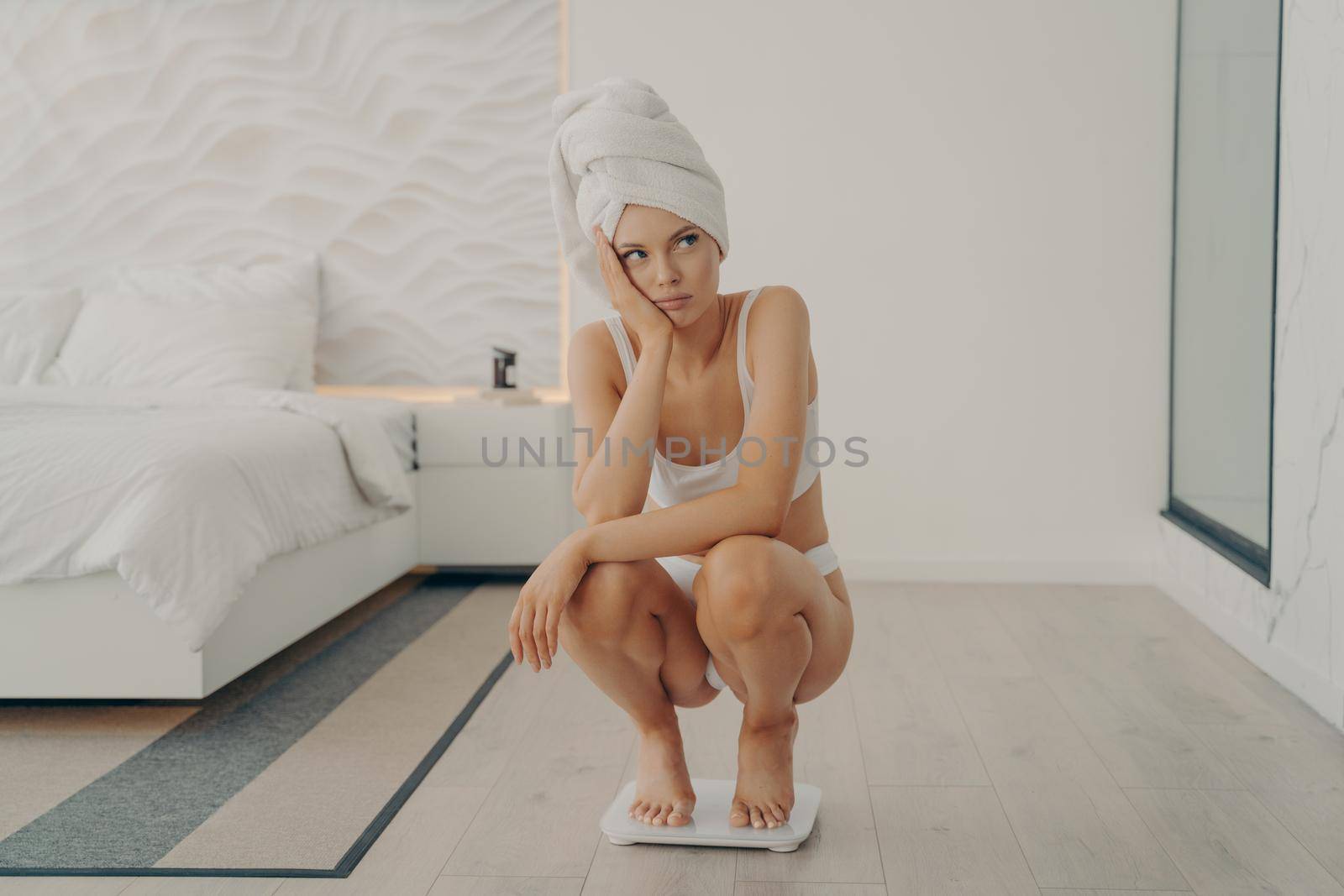 Frustrated sad young european girl squats on scales in bedroom after shower, gaining and not losing excess weight, having hard time to slim. Weight loss problem concept