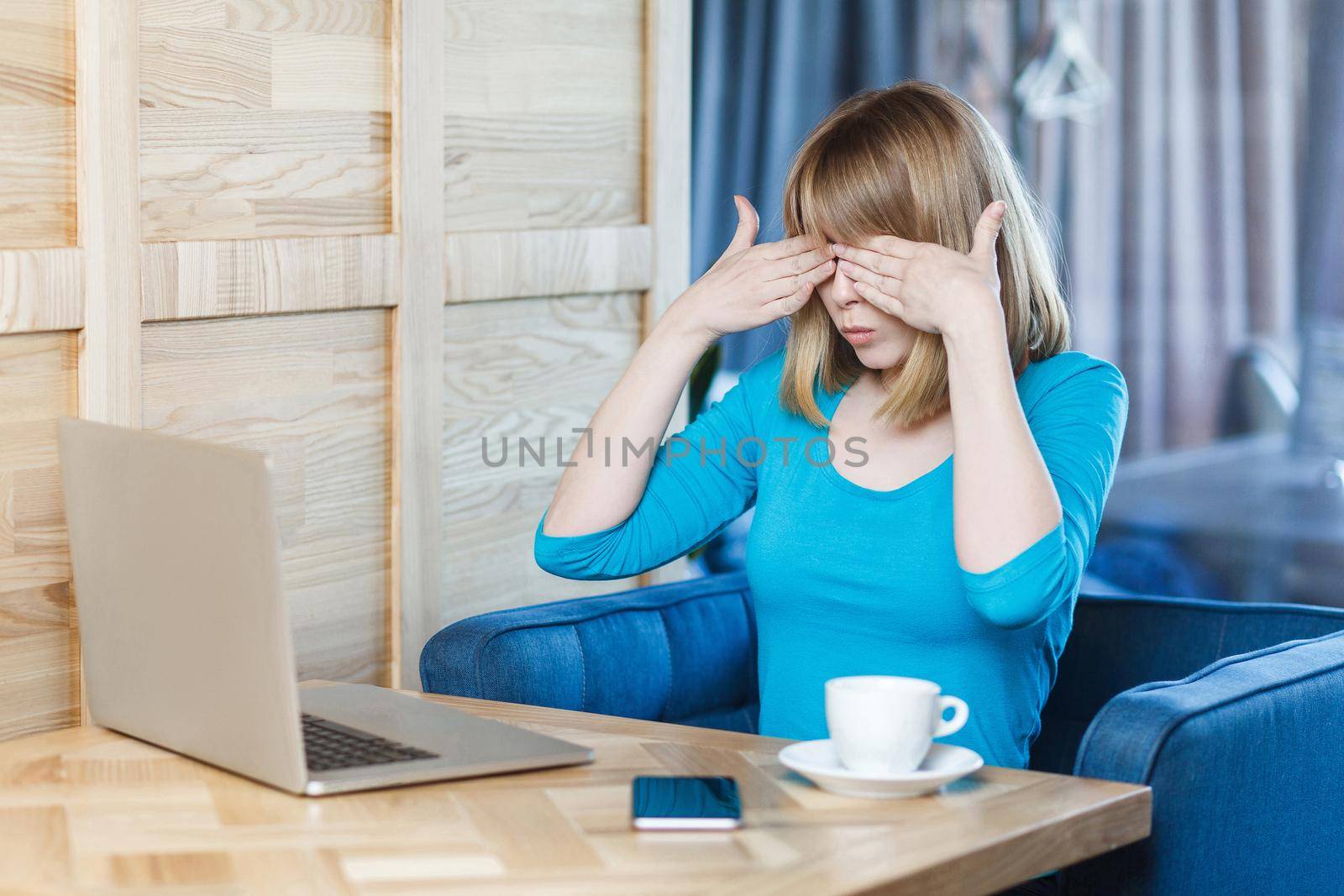 Side view portrait of emotional scared young businesswoman in blue t-shirt are sitting in cafe and covering the eyes to herself with hand cause made big mistake in the laptop. Indoor, inside