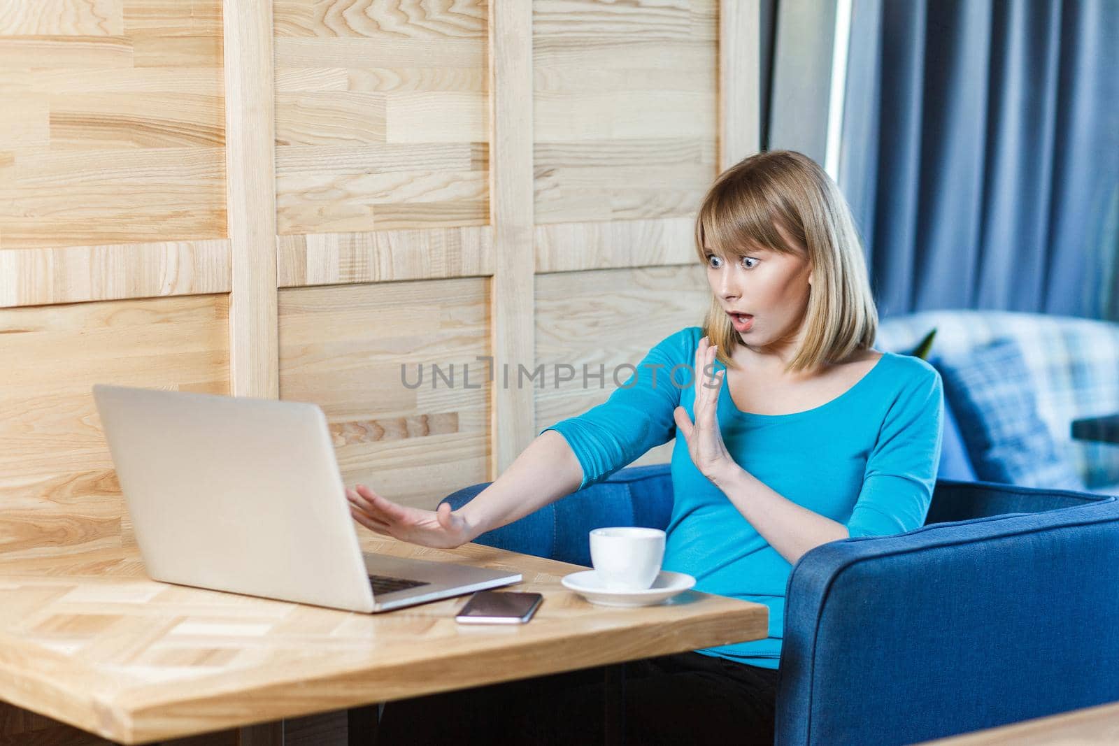 Stop! Side view portrait of emotional scared young girl freelancer in blue blouse are sitting in cafe and screaming to the laptope cause made big mistake with raised arms and stressed face. Indoor