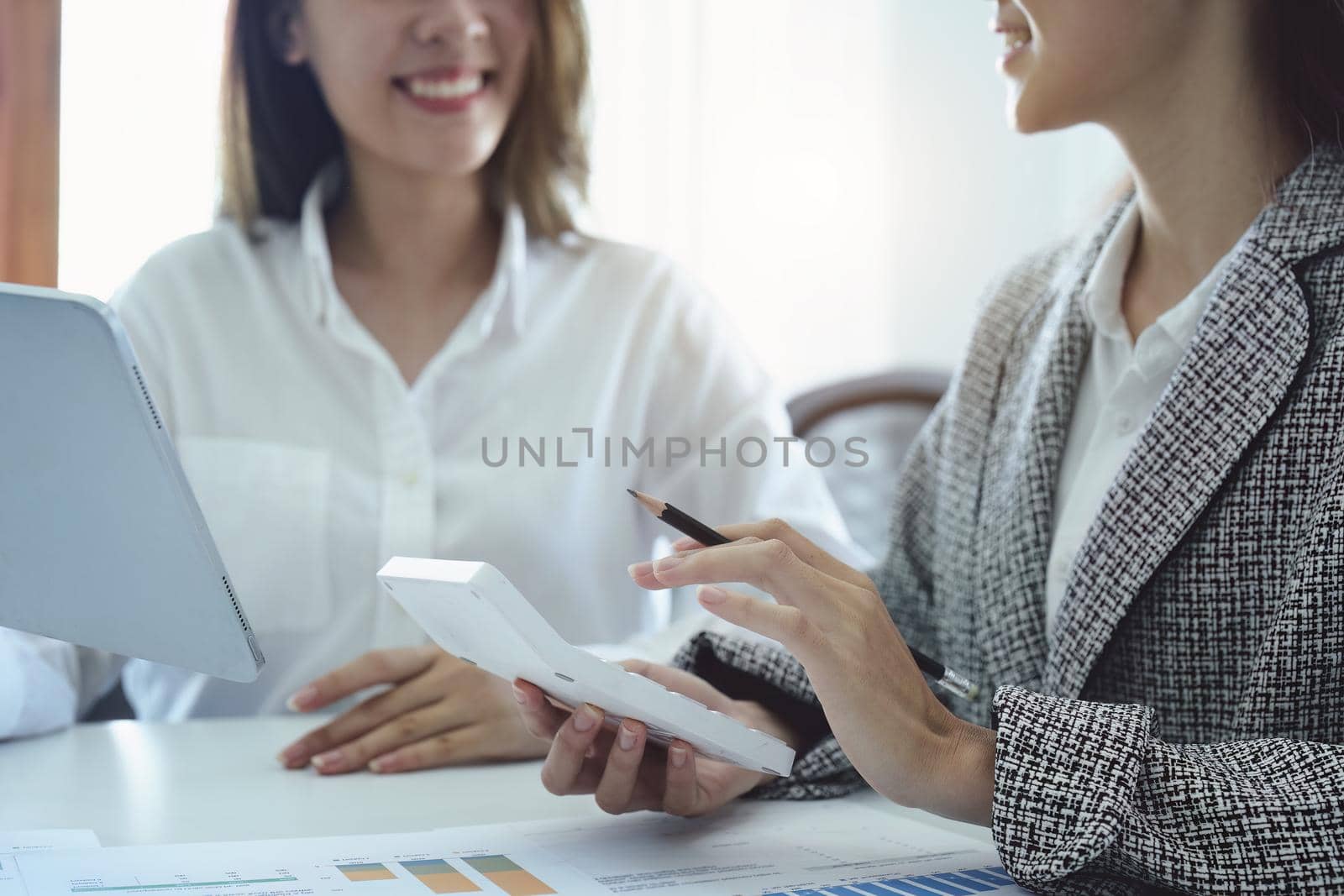 two young Asian businesswoman using their tablet computers with calculators and documents to plan marketing strategies and investments to profit from their clients by Manastrong