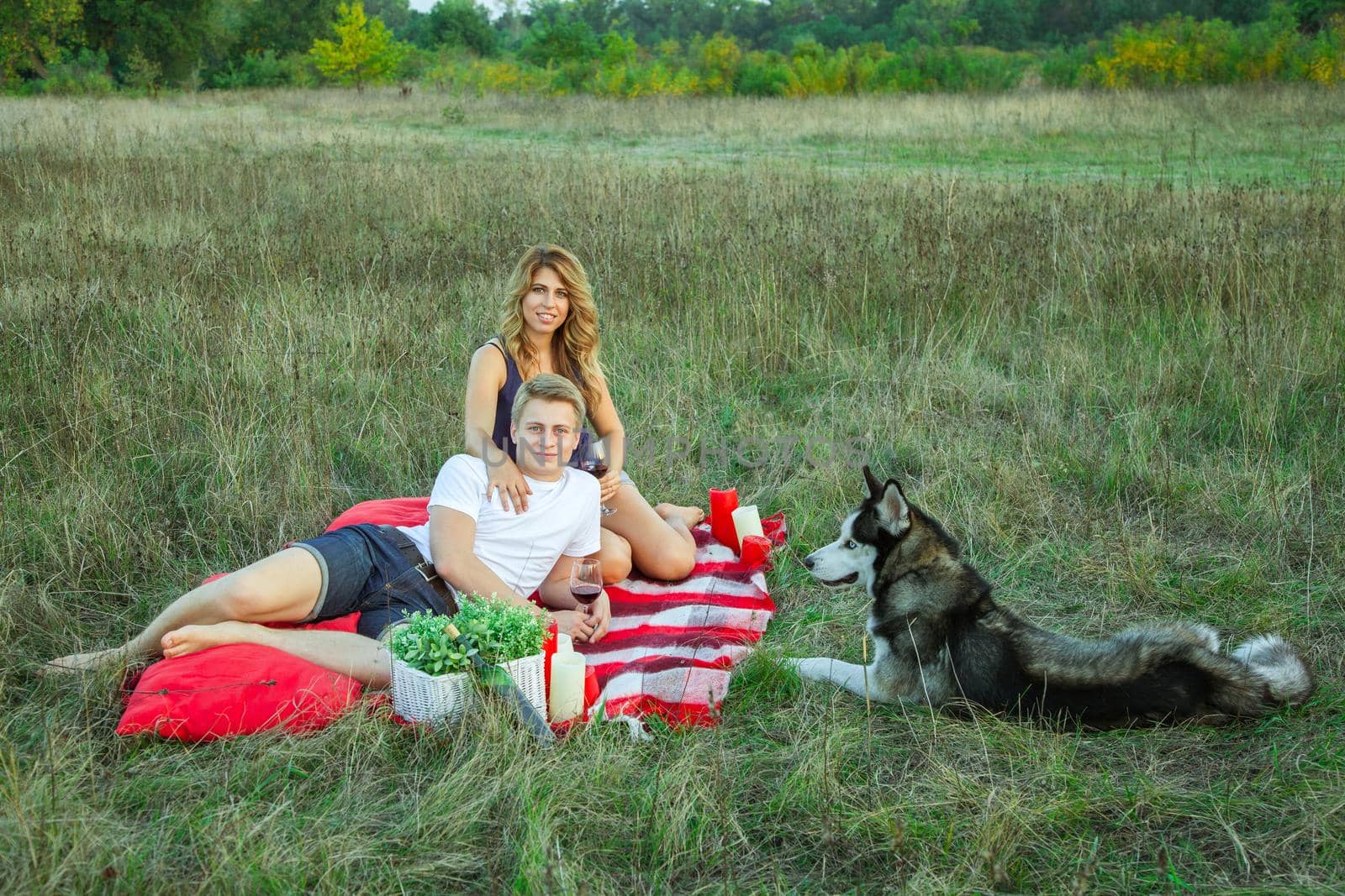 Beautiful young happy loving couple on picnic lying down on plaid with their dog in field on sunny summer day enjoying, holding and drinking wine and resting. looking at camera and smiling.