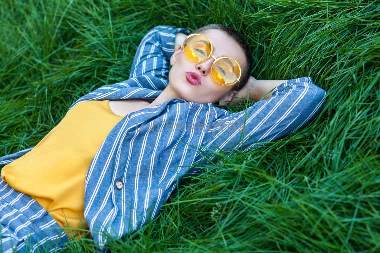 Portrait of cute young girl with short hair in casual blue striped suit, yellow shirt, glasses lying down on green grass, resting, holding hand behind head, looking at camera, kissing. summertime shot