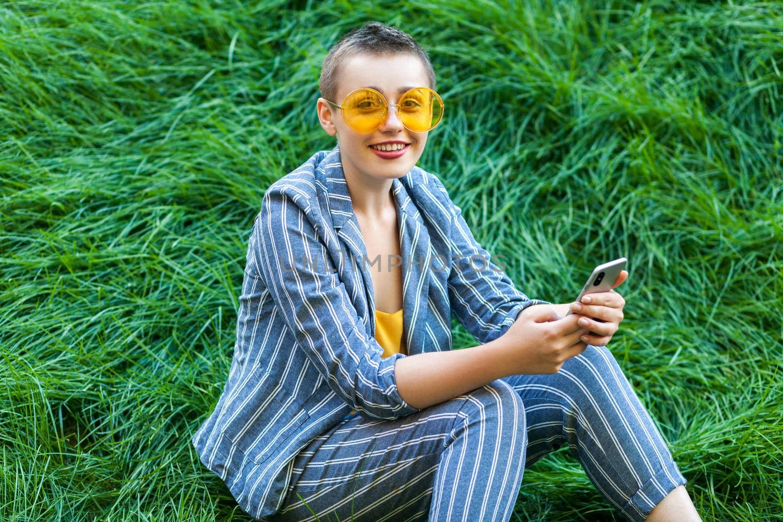 Portrait of pretty young short hair woman in casual blue striped suit, yellow glasses sitting on grass holding her smart phone mobile and looking at camera with toothy smile. outdoor summertime shot.