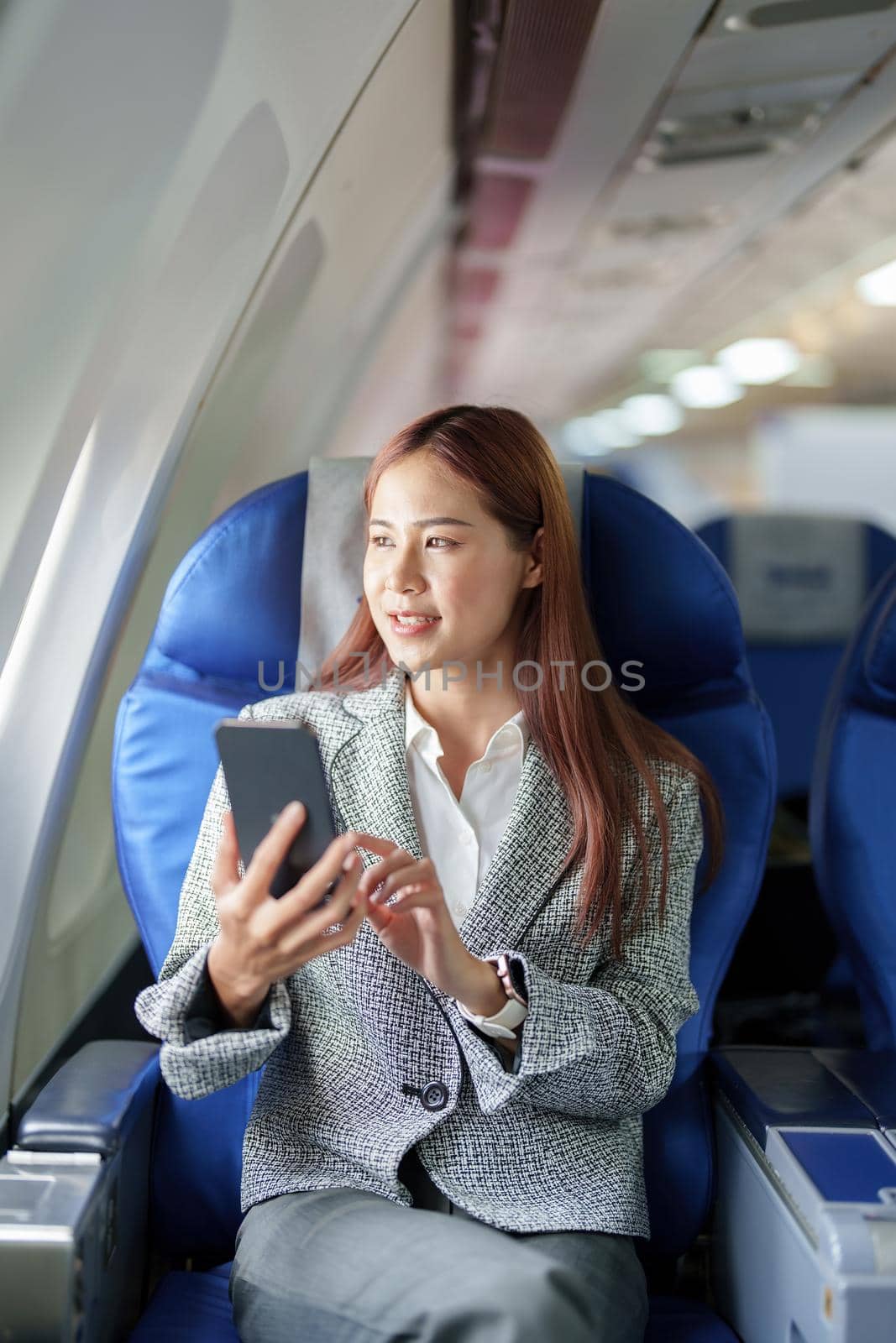 A portrait of a smiling Asian businesswoman using her phone while on a plane by Manastrong