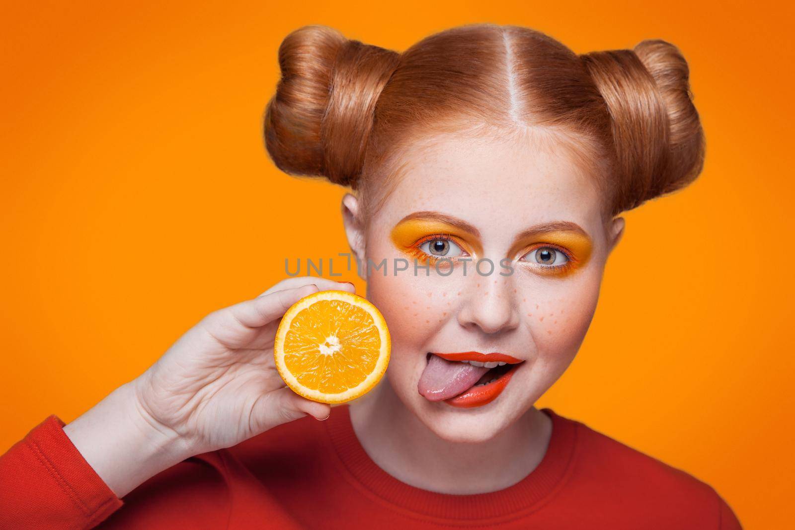 Young beautiful funny fashion model with orange slice on orange background. with orange makeup and hairstyle and freckles. studio shot, looking at camera and shows her tongue.
