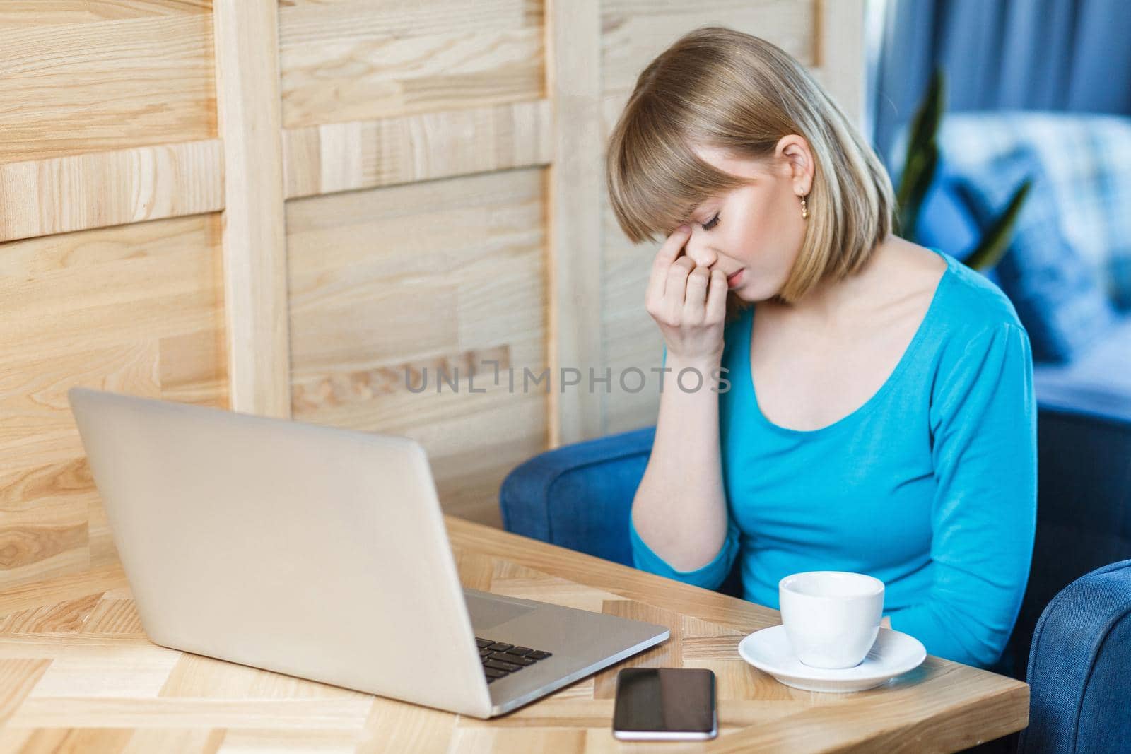 Side view portrait of sad young girl freelancer with blonde bob haircut hair in blue t-shirt are sitting in cafe and working on laptop,closed eyes and trying to stop crying hold fingers on the eyes