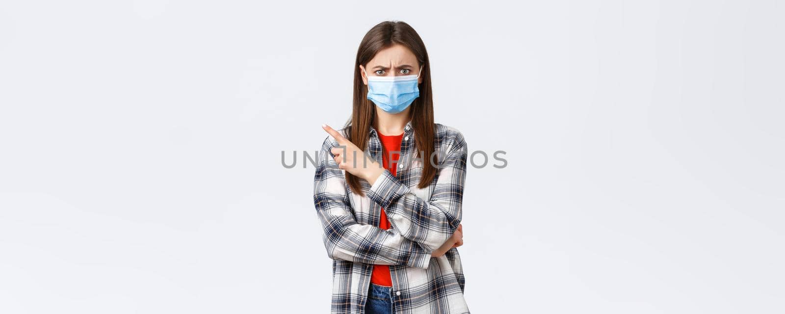 Coronavirus outbreak, leisure on quarantine, social distancing and emotions concept. Angry and disappointed young female in medical mask, pointing finger upper left corner, frowning mad.