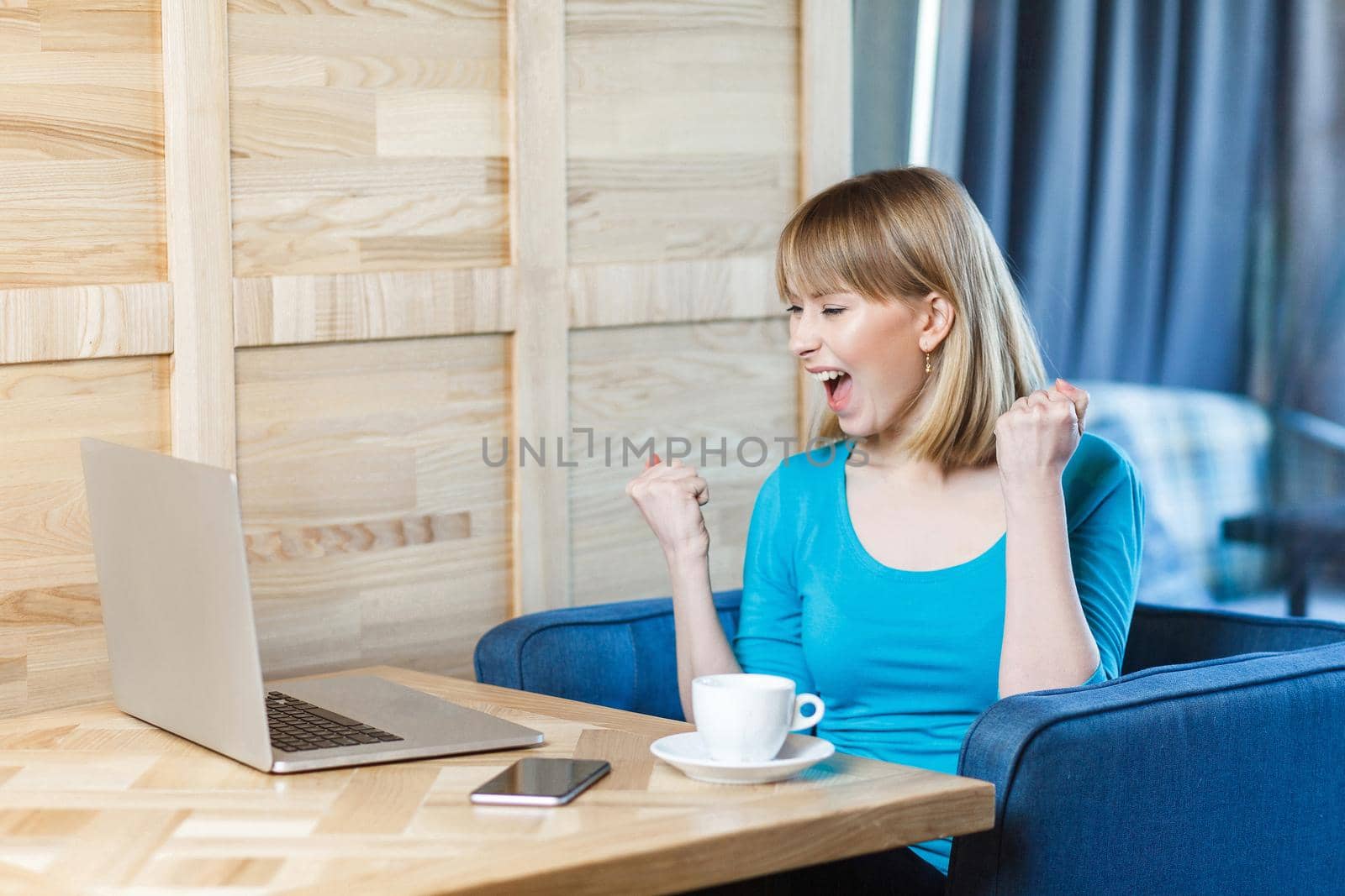 Side view portrait of beautiful amazed satisfied positive young girl freelancer with blonde hair in blue t-shirt are sitting in cafe and watching online reality show with toothy smile and triumphing