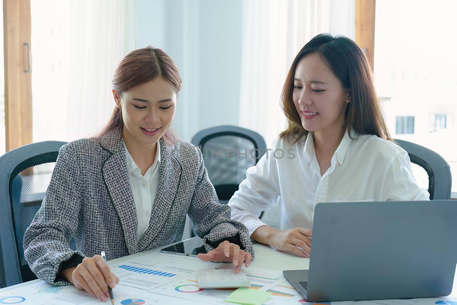 two young Asian businesswoman using their tablet computers with calculators and documents to plan marketing strategies and investments to profit from their clients.