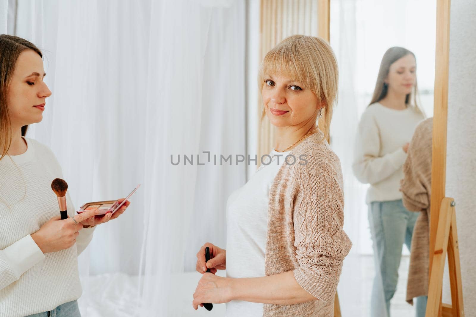 Beautiful smiling young caucasian mature senior mother mom and adult daughter doing makeup, standing near mirror, and looking at reflection in living room home. Family with two women relax together.