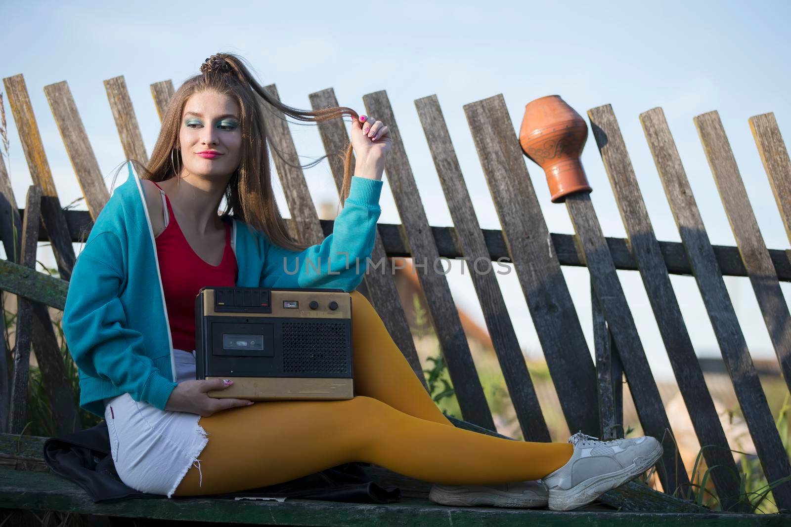 A beautiful country girl in bright clothes sits on a wooden bench with a cassette recorder. Woman in the style of the 90s.