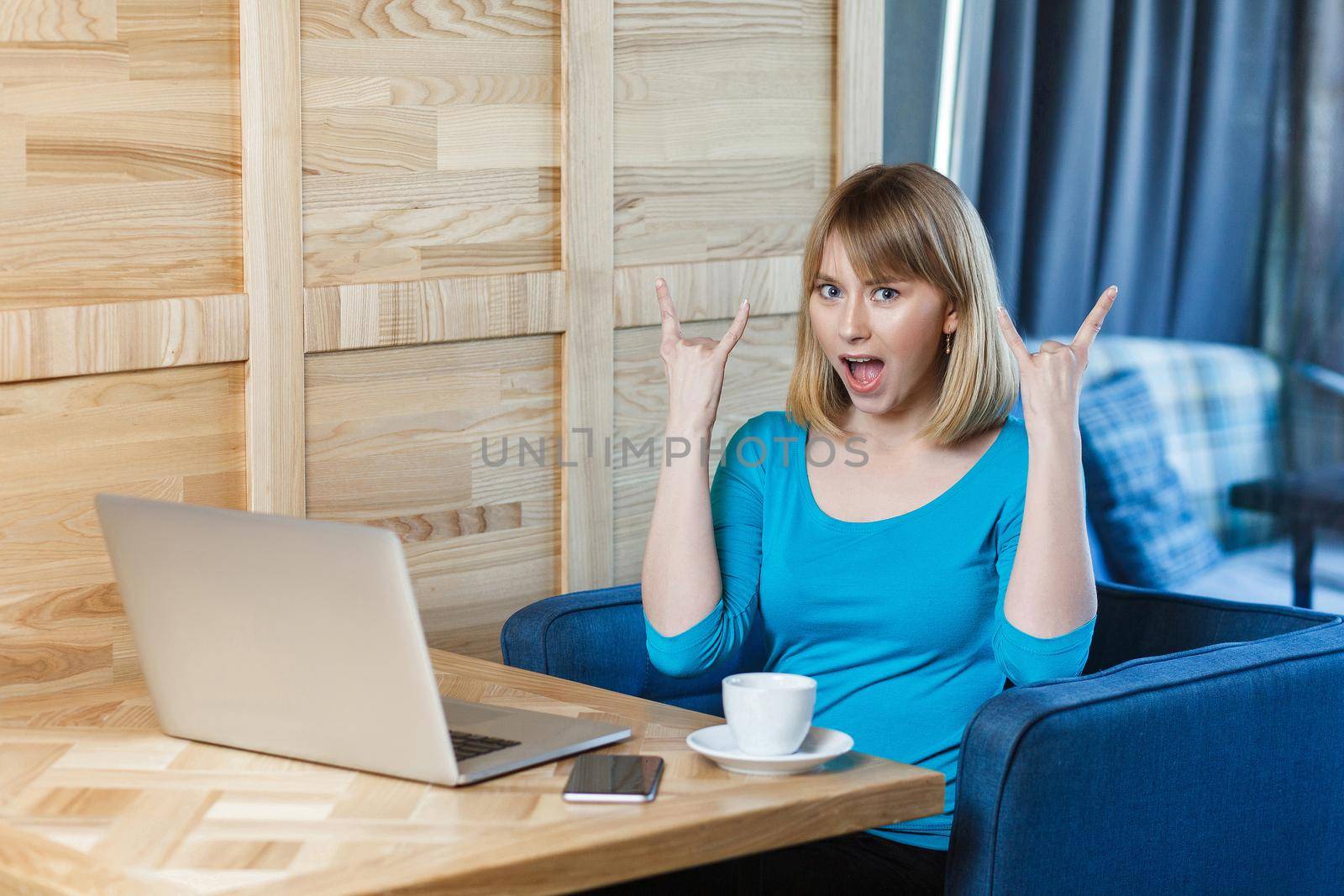 Portrait of attractive successfull young girl freelancer with blonde hair in blue blouse are sitting in cafe and working on laptop with toothy smile and showing rock sign, looking at camera. indoor