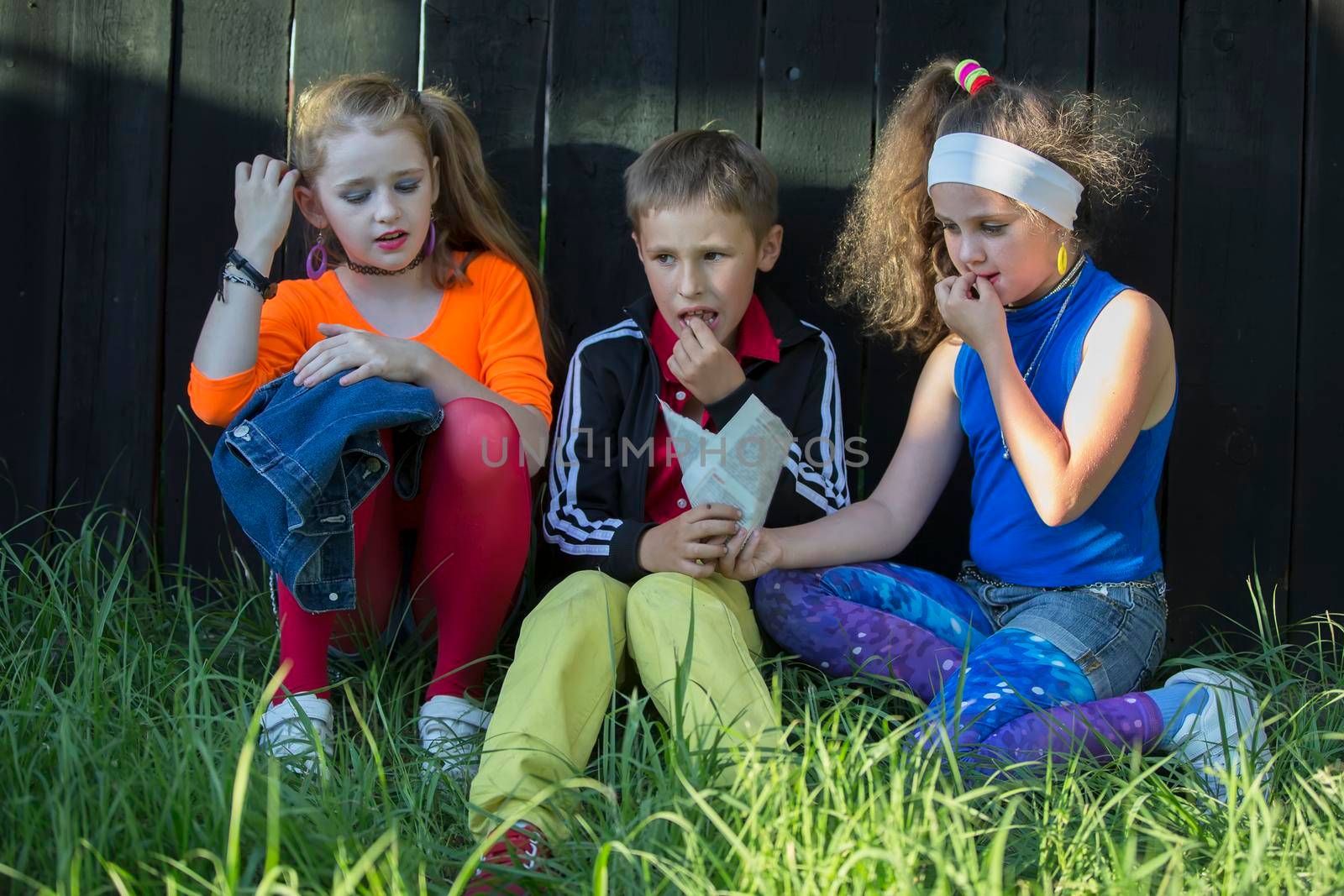 Cheerful kids, a boy and two cool girls sit on the grass near a wooden fence. by Sviatlana