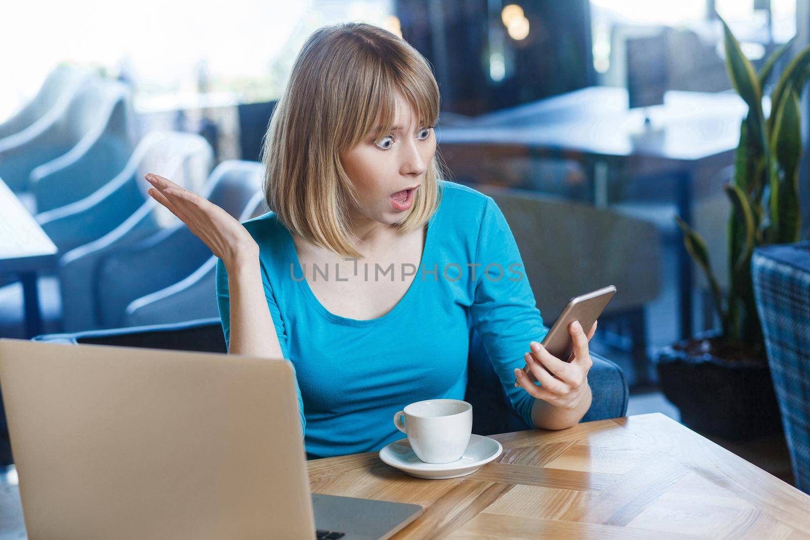 Top view portrait of young shocked blonde woman in blue t-shirt, sitting in cafe and holding her mobile smart phone and looking with amazed face and big eyes with raised hand. indoor studio shot.