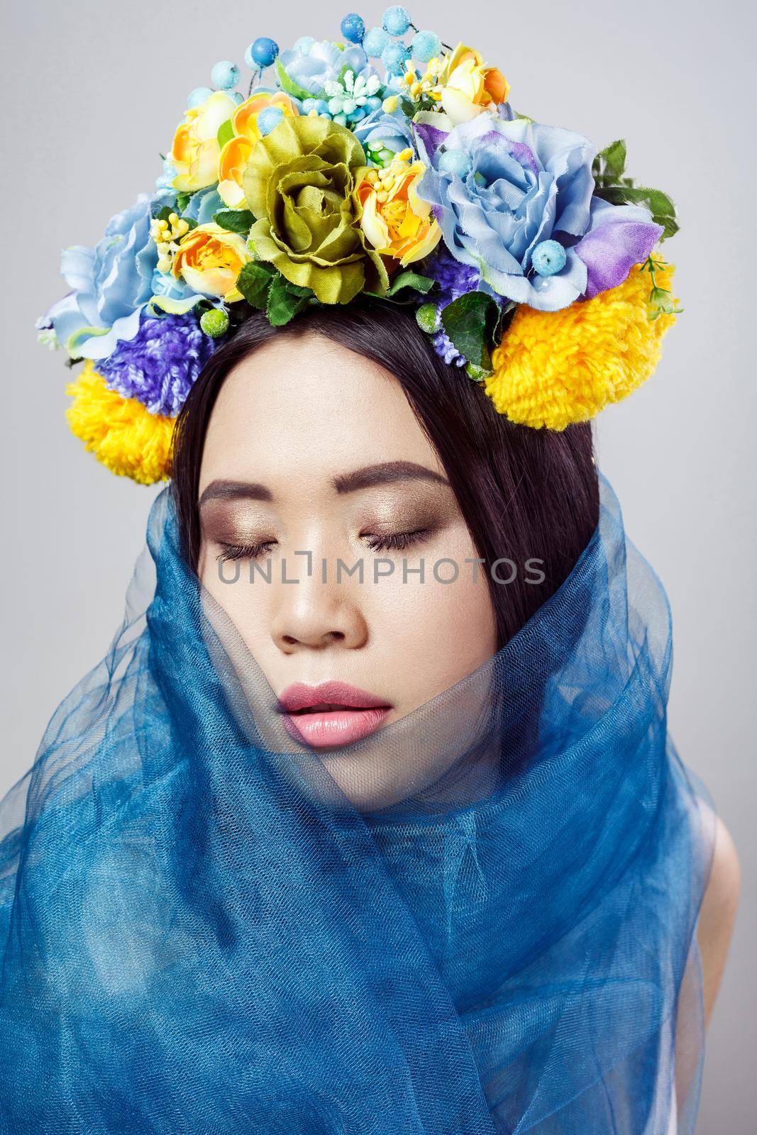 portrait of attractive asian woman with floral hat and blue veil posing with closed eyes on light grey background . indoor studio shot.