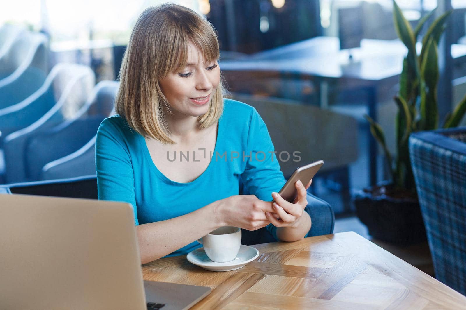 Top view portrait of young happy blonde woman in blue t-shirt, sitting in cafe and holding her mobile smart phone and texting. indoor studio shot.