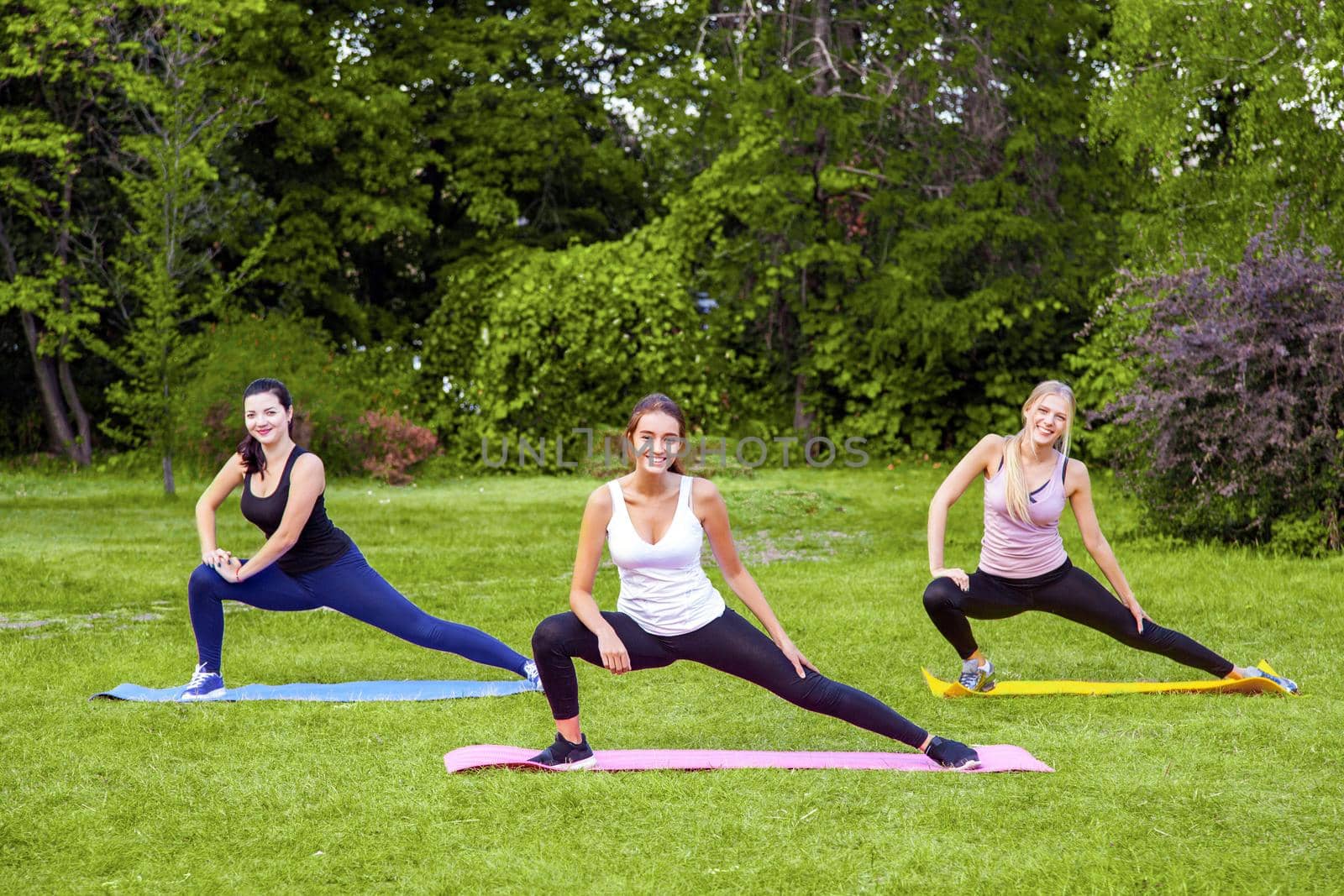 Group of beautiful healthy slimy woman doing exersices on mat in the green grass in the park, streching legs, looking at camera with toothy smile. by Khosro1