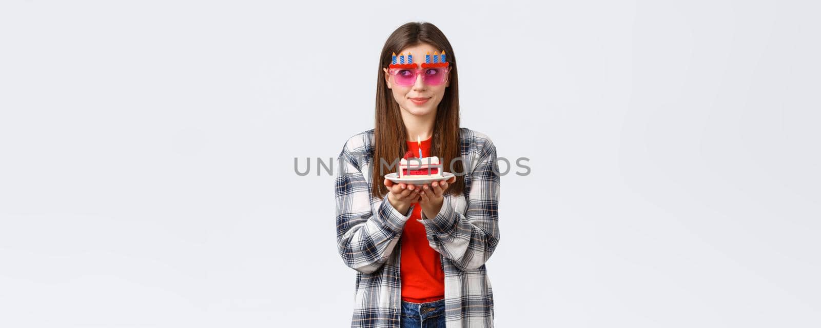 People lifestyle, holidays and celebration, emotions concept. Cute and silly birthday girl in funny glasses, look away think of wish as blowing lit candle on b-day cake, smiling excited.